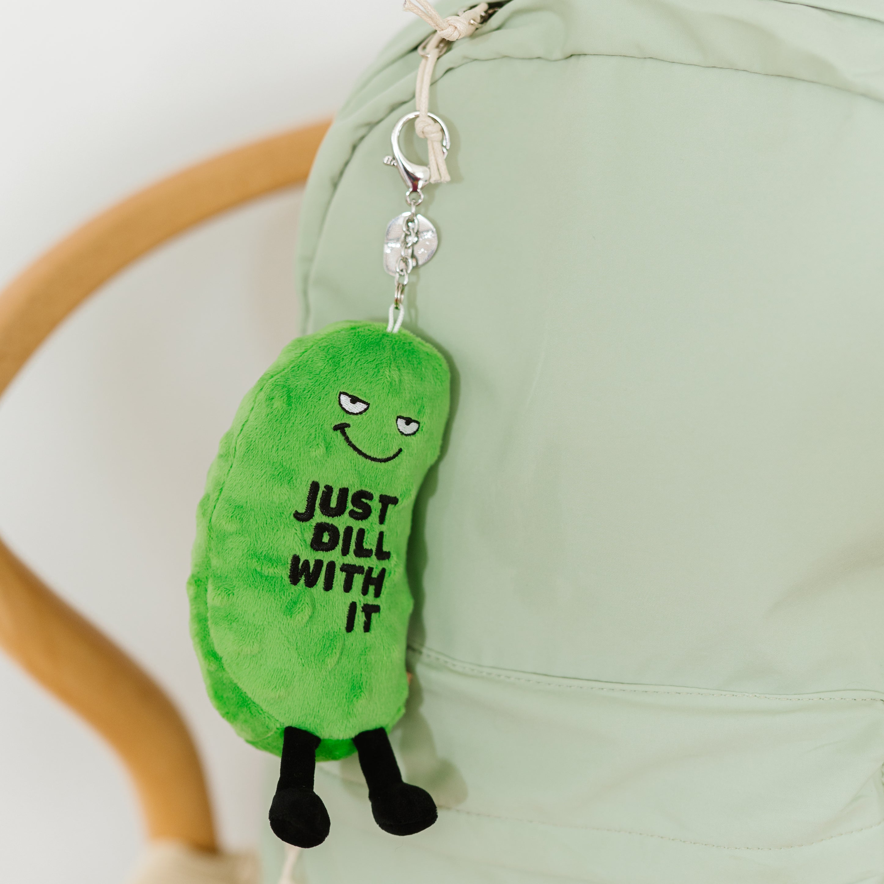 A plush pickle charm with the text 'JUST DILL WITH IT' printed on it hanging off a backpack.