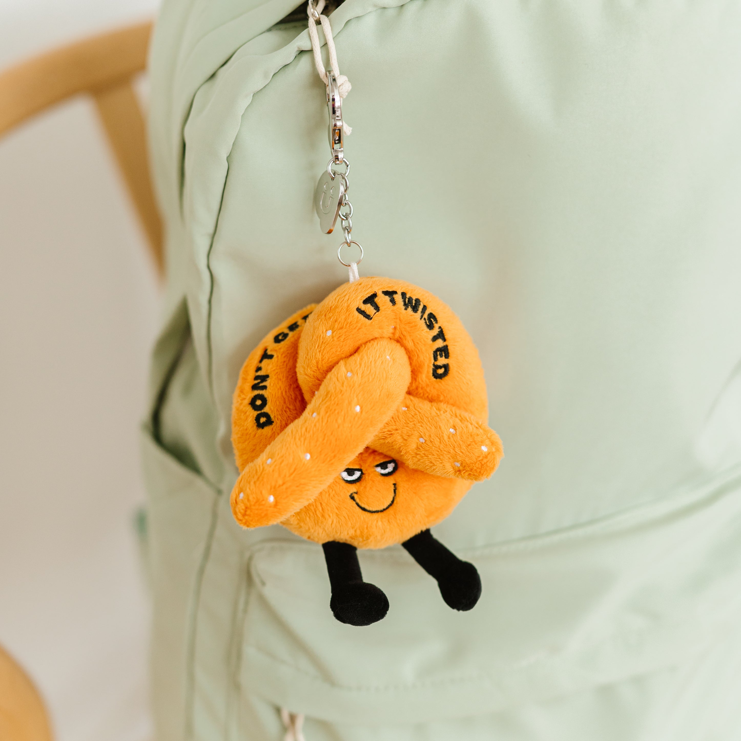 A plush bag charm shaped like a pretzel with a smiley face and the text 'DON'T GET IT TWISTED' printed on it hanging off a backpack. 