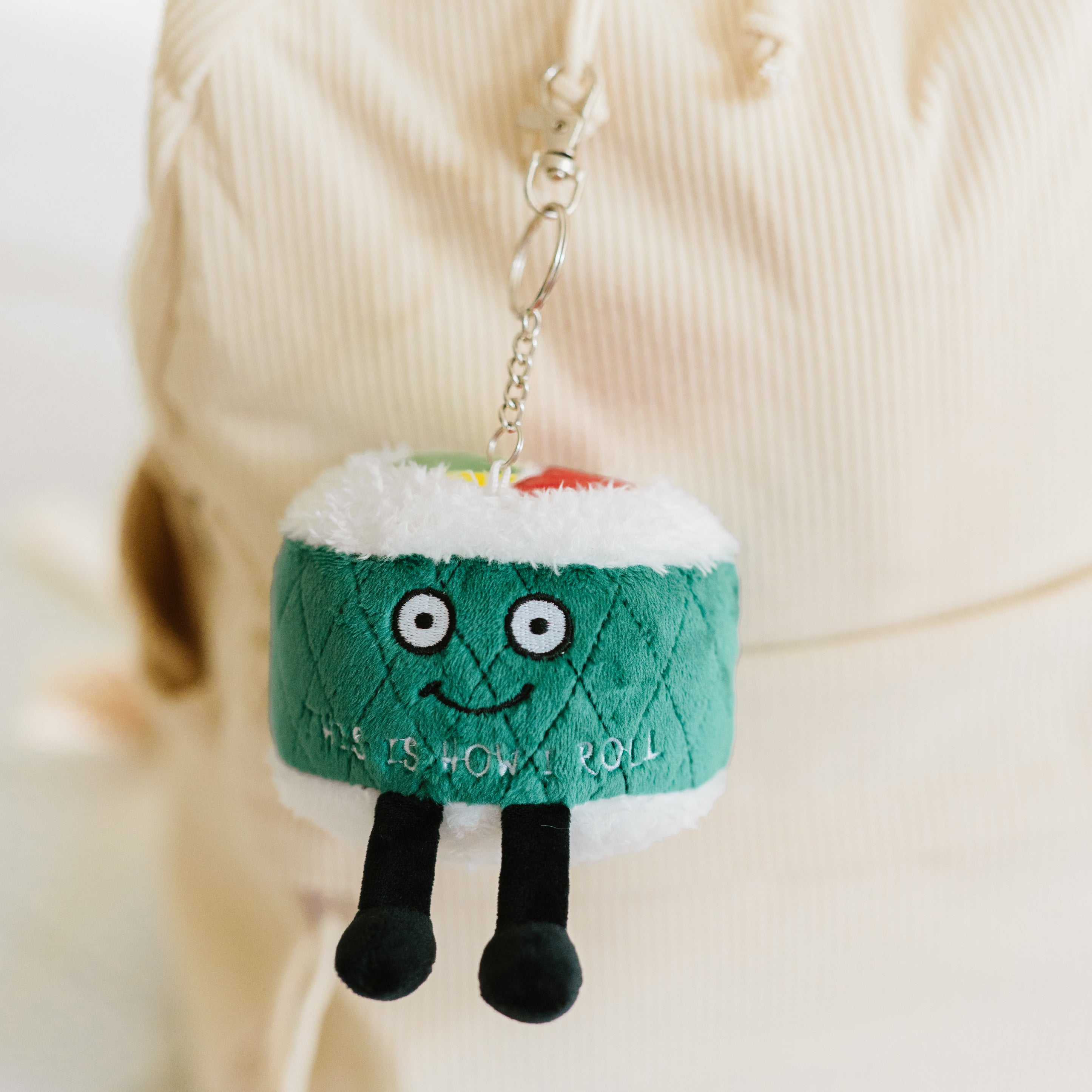 A plush charm designed to look like a piece of sushi with a smiling face, eyes, and dangly legs. The charm has the text 'THIS IS HOW I ROLL' written on it hanging on a backpack.