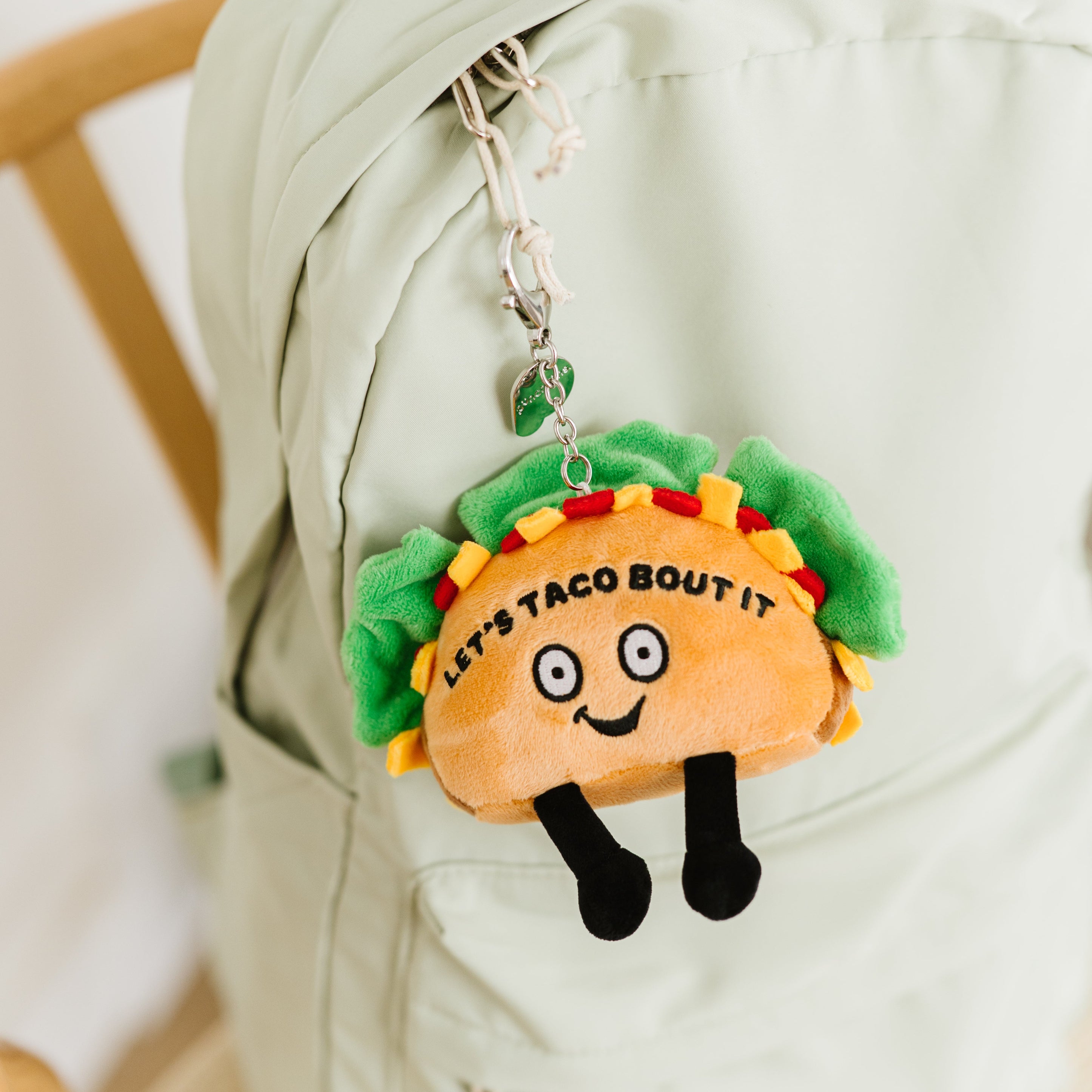 A plush bag charm designed to look like a taco with eyes and dangly legs with text that says 'LET'S TACO 'BOUT IT' hanging off a backpack.