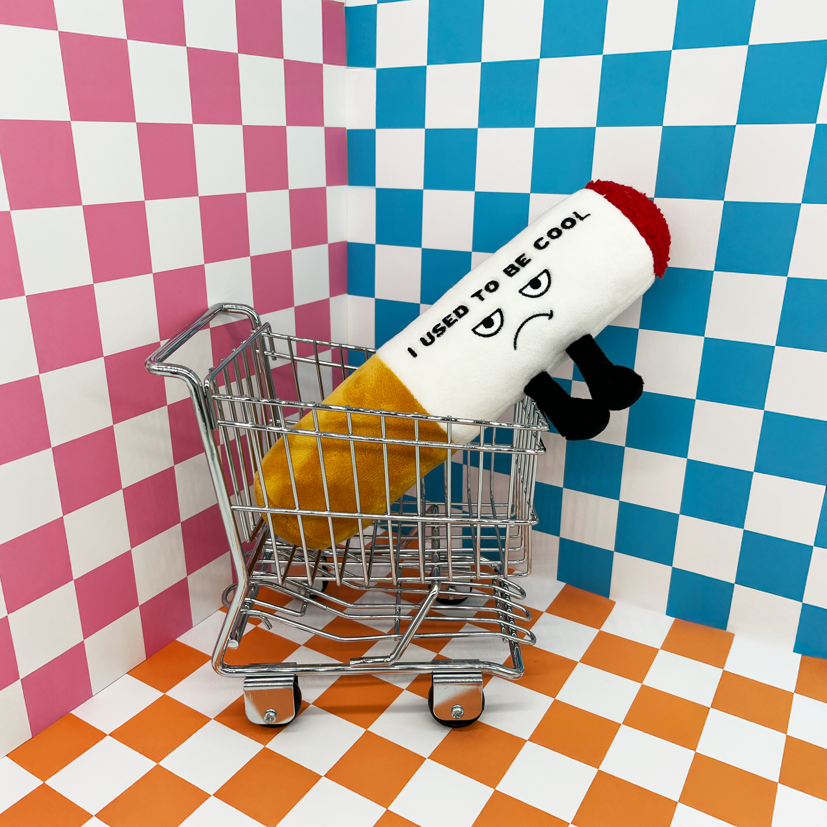 A plush cigarette in a mini shopping cart on a checkered background. 