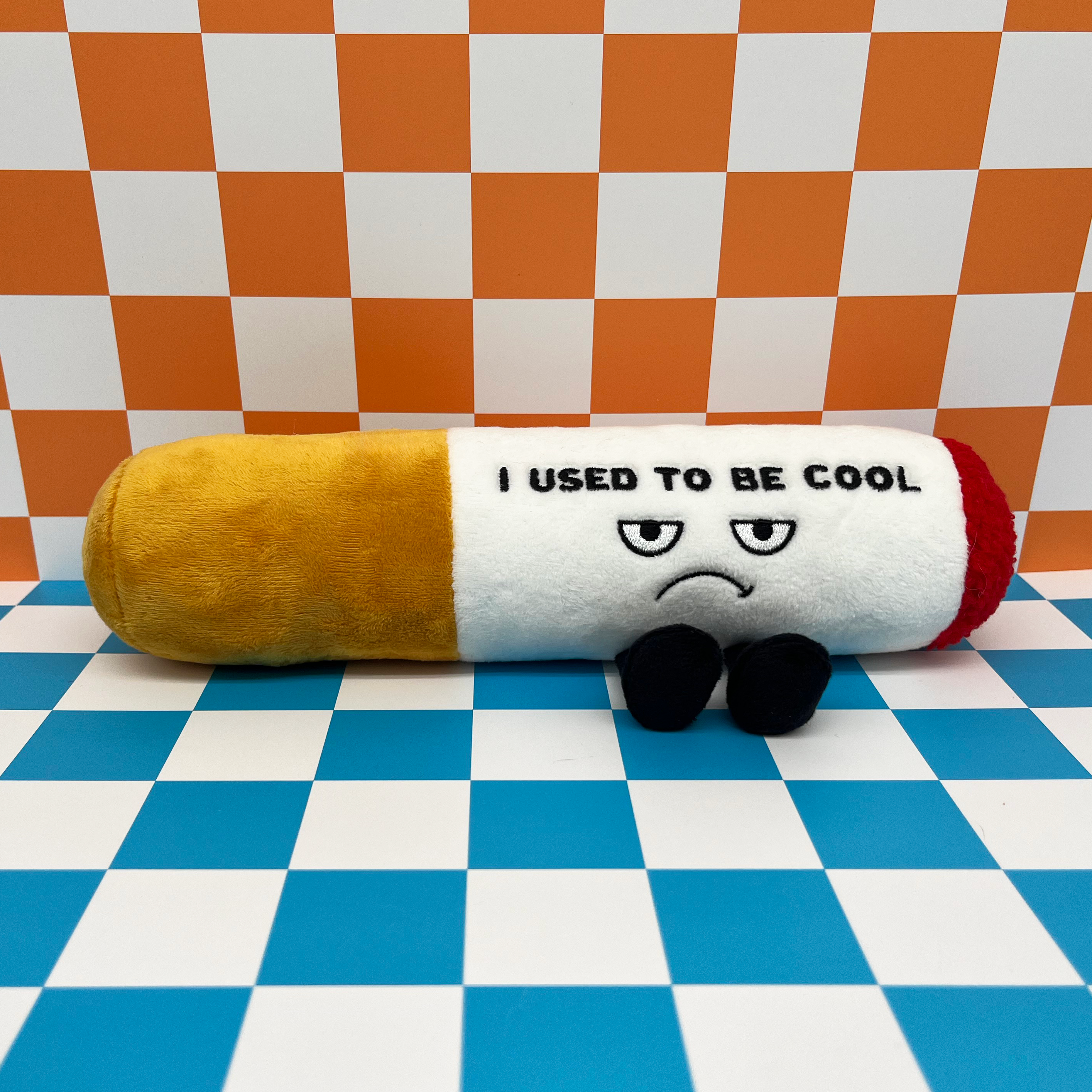 A plush cigarette with an upset face expression and the text 'I USED TO BE COOL' on it placed on a checkered backround.