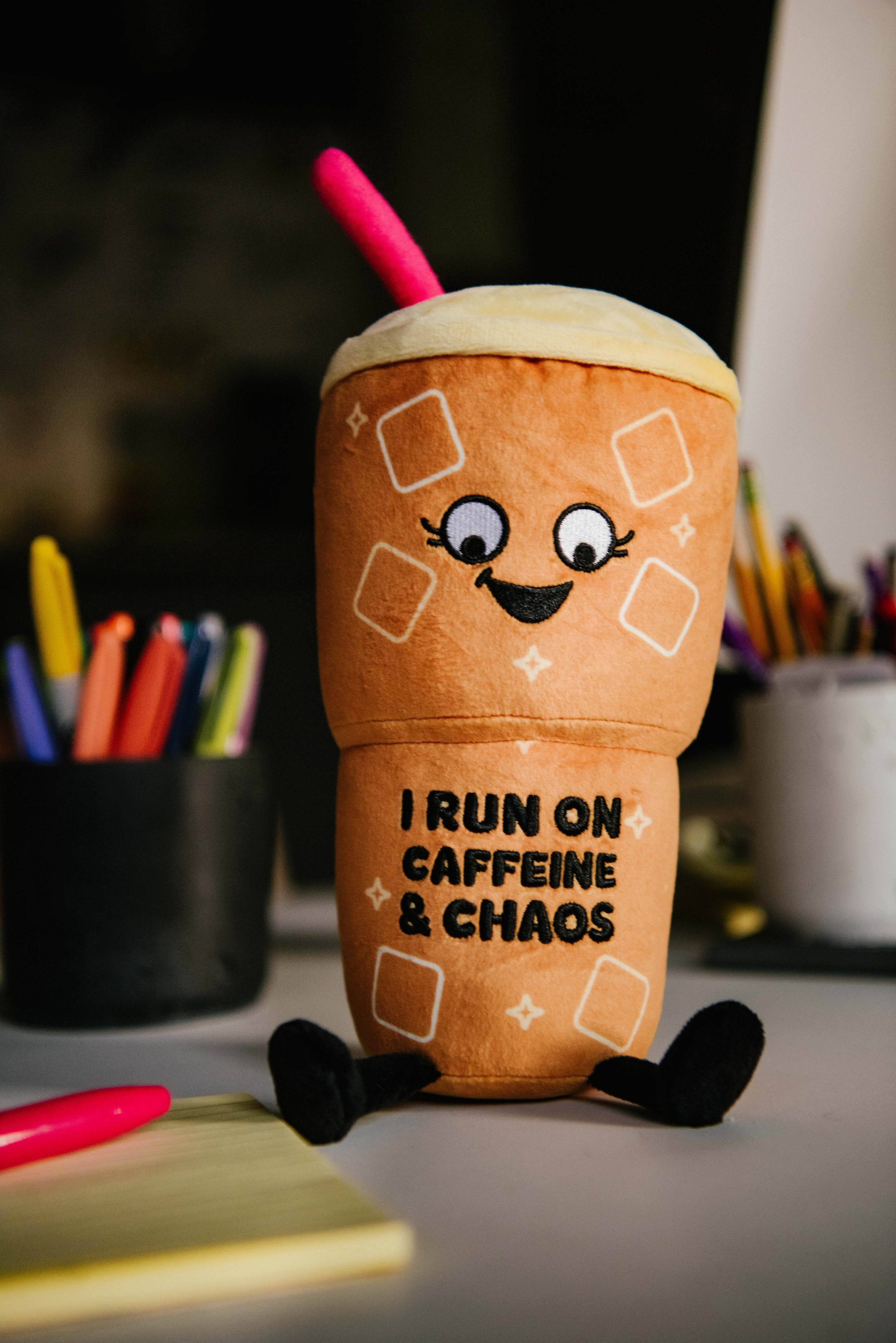 A plush designed to look like an iced coffee cup with the text 'I RUN ON CAFFEINE & CHAOS' on a desk surrounded by office supplies.