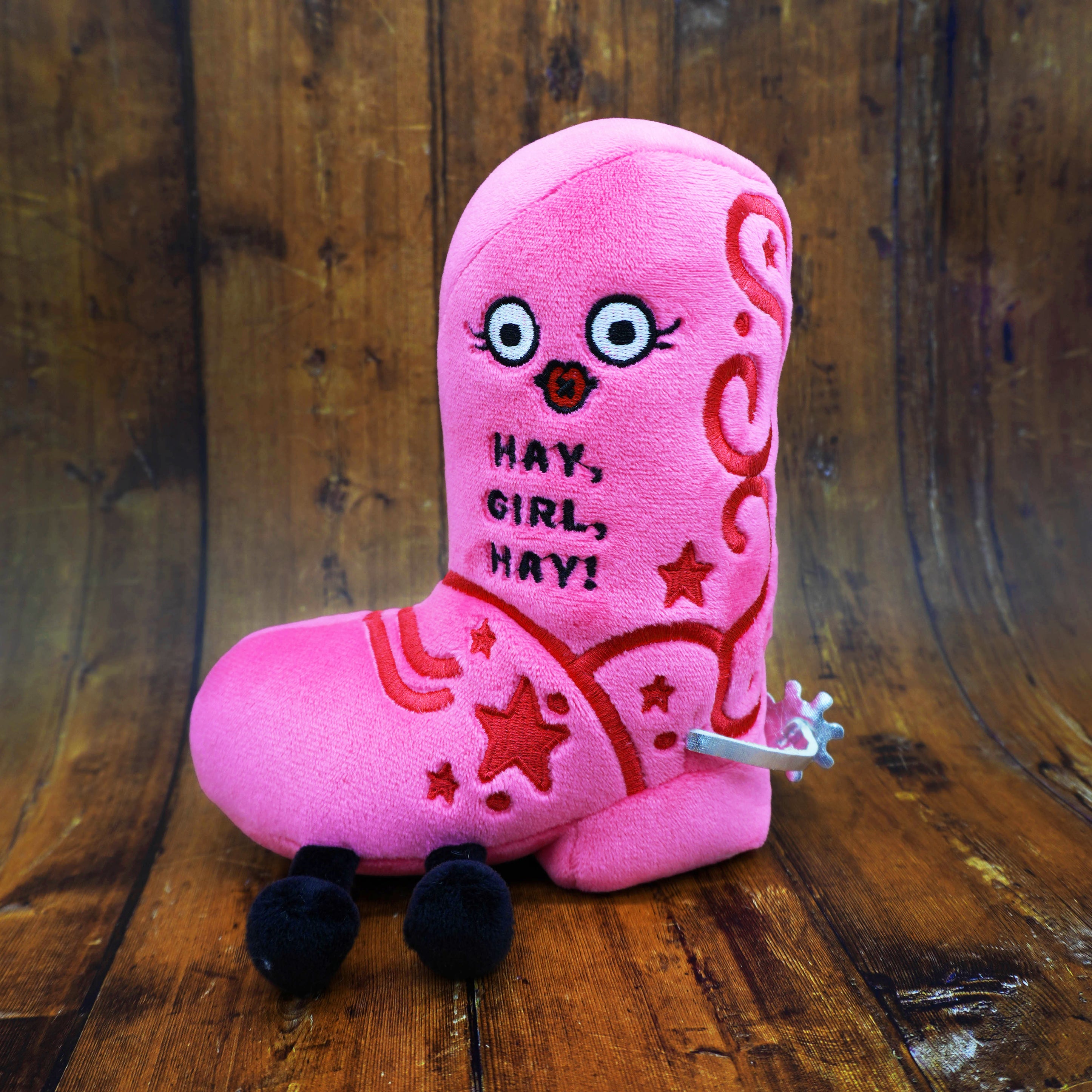 Pink plush toy cowgirl boot with cartoon face and text that reads, "Hay, girl, hay" on a wooden surface. 