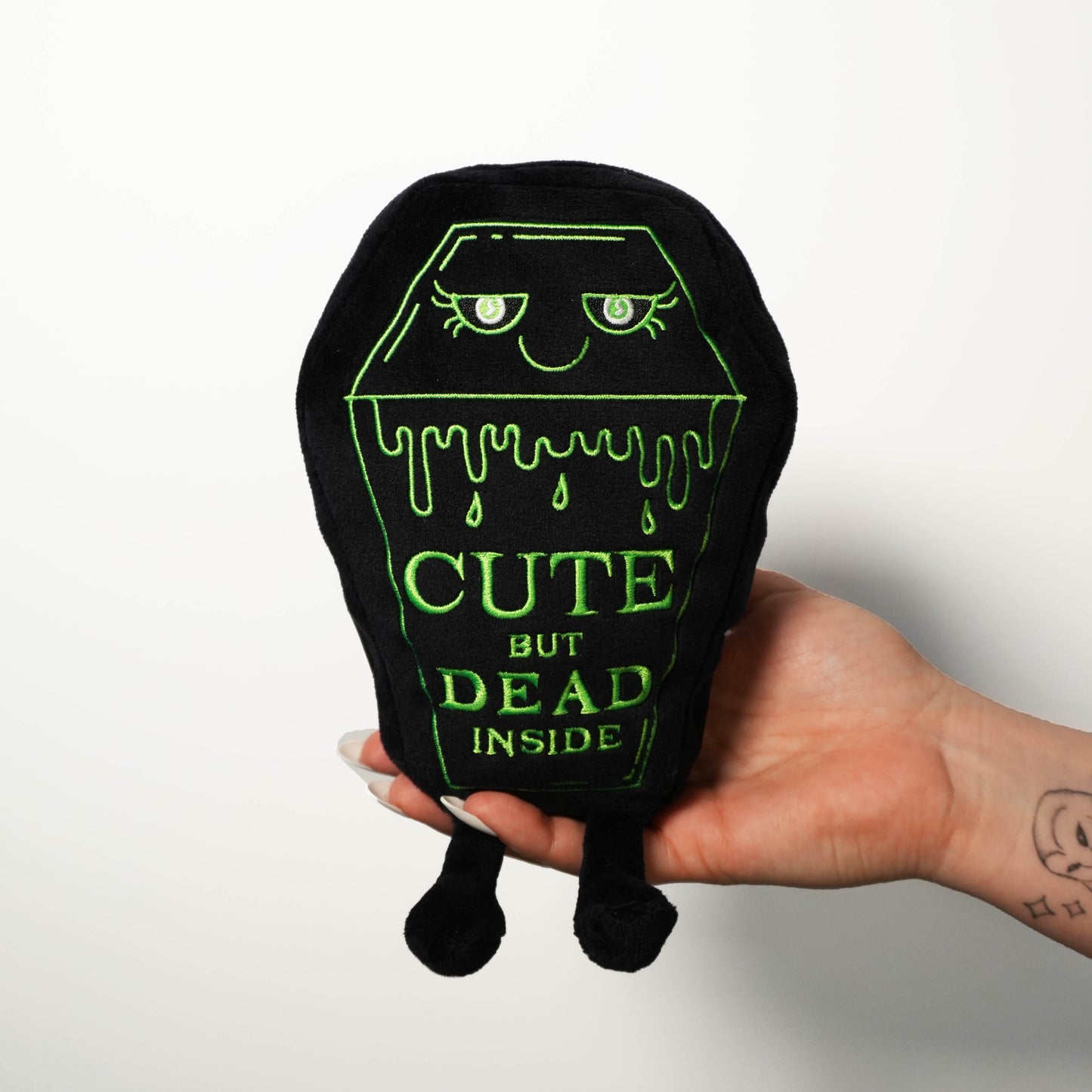 Black plush toy with green text 'Cute but dead inside' held by a hand on a white background