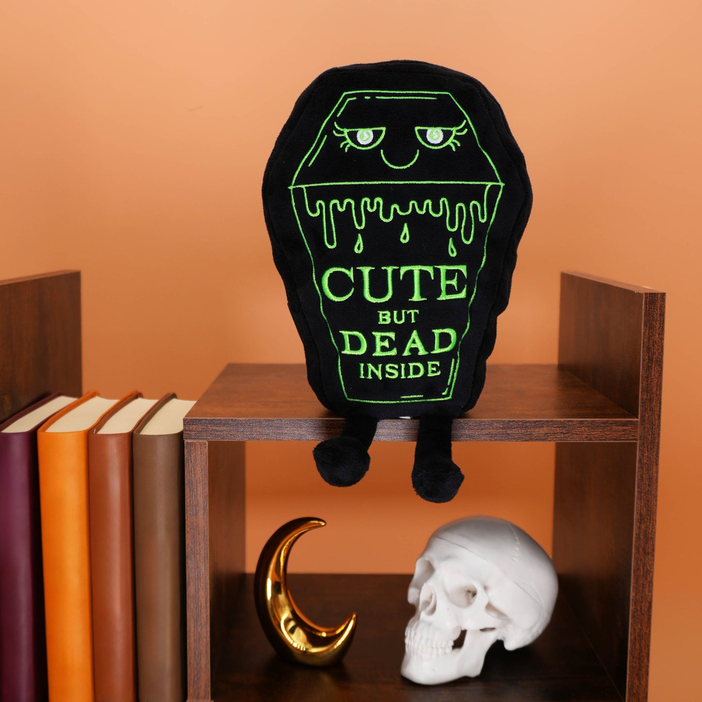 Black plush toy with green text 'Cute but dead inside' on a wooden shelf with books and a skull.