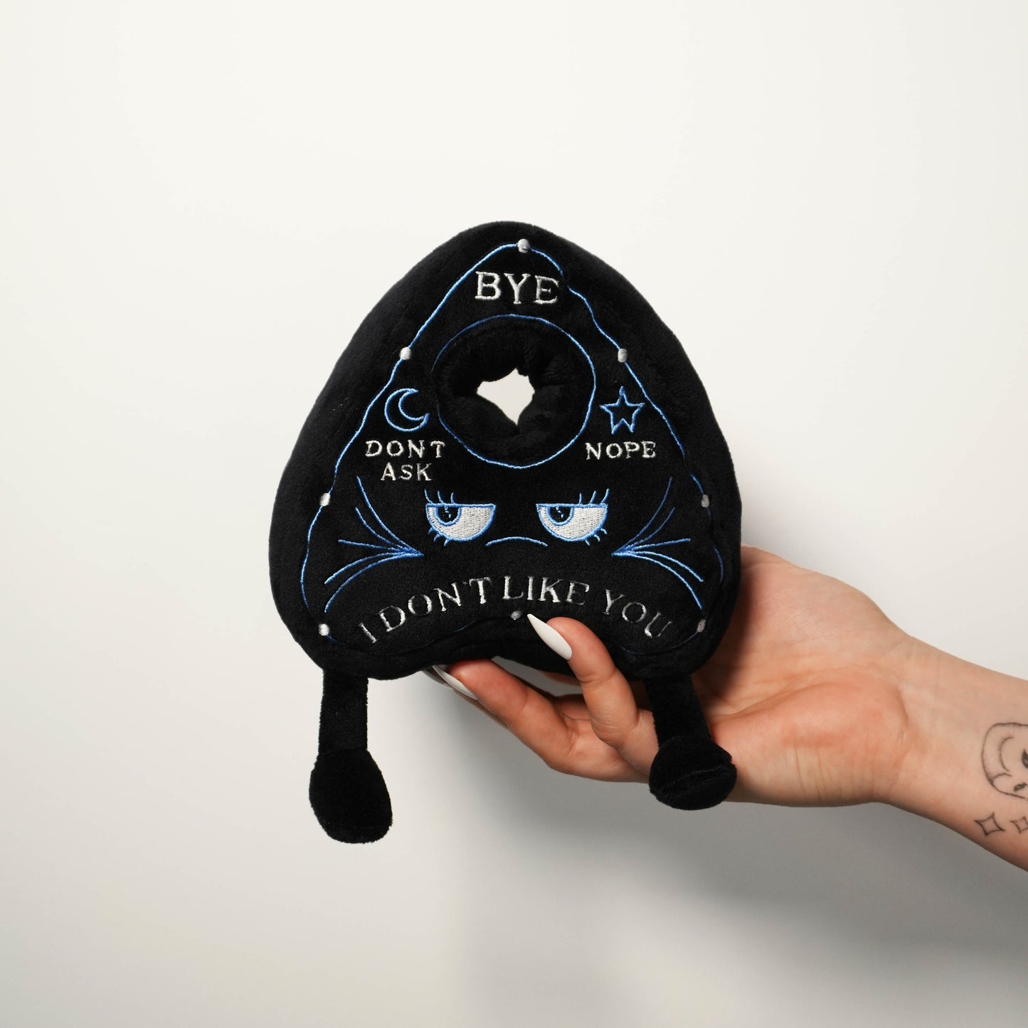 Hand holding a black planchette plush toy with text and eyes on a white background