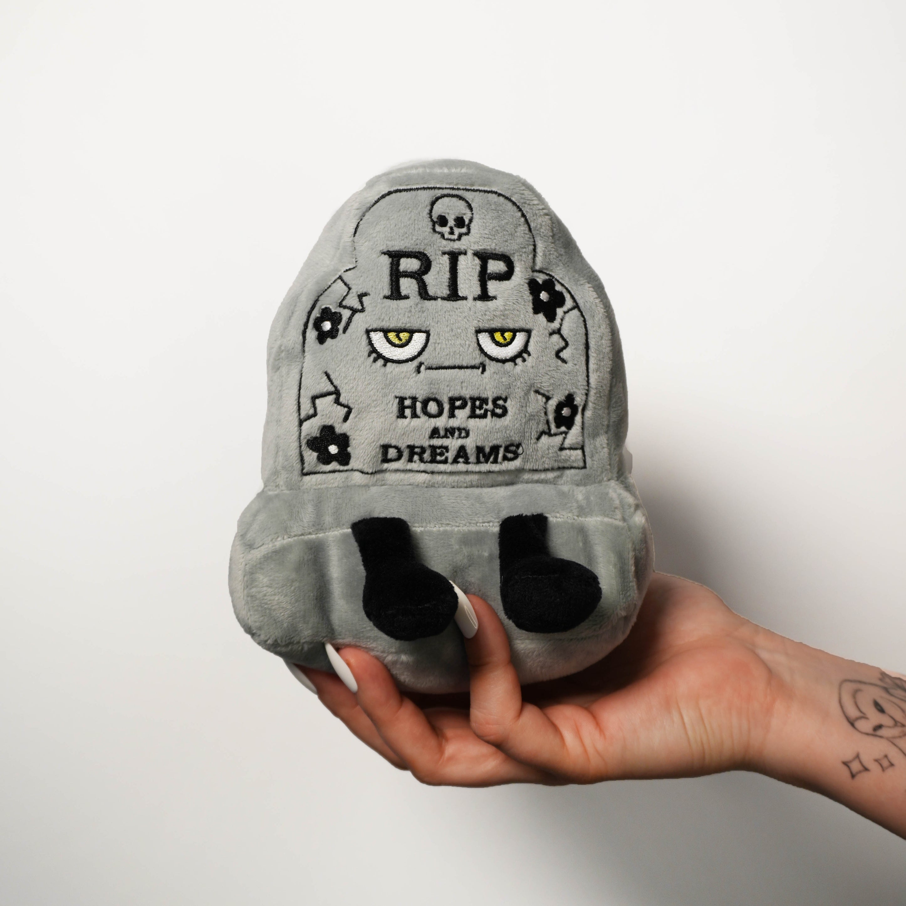 Gray plush toy resembling a headstone with "RIP Hopes and Dreams" text, held in a hand against a white background.