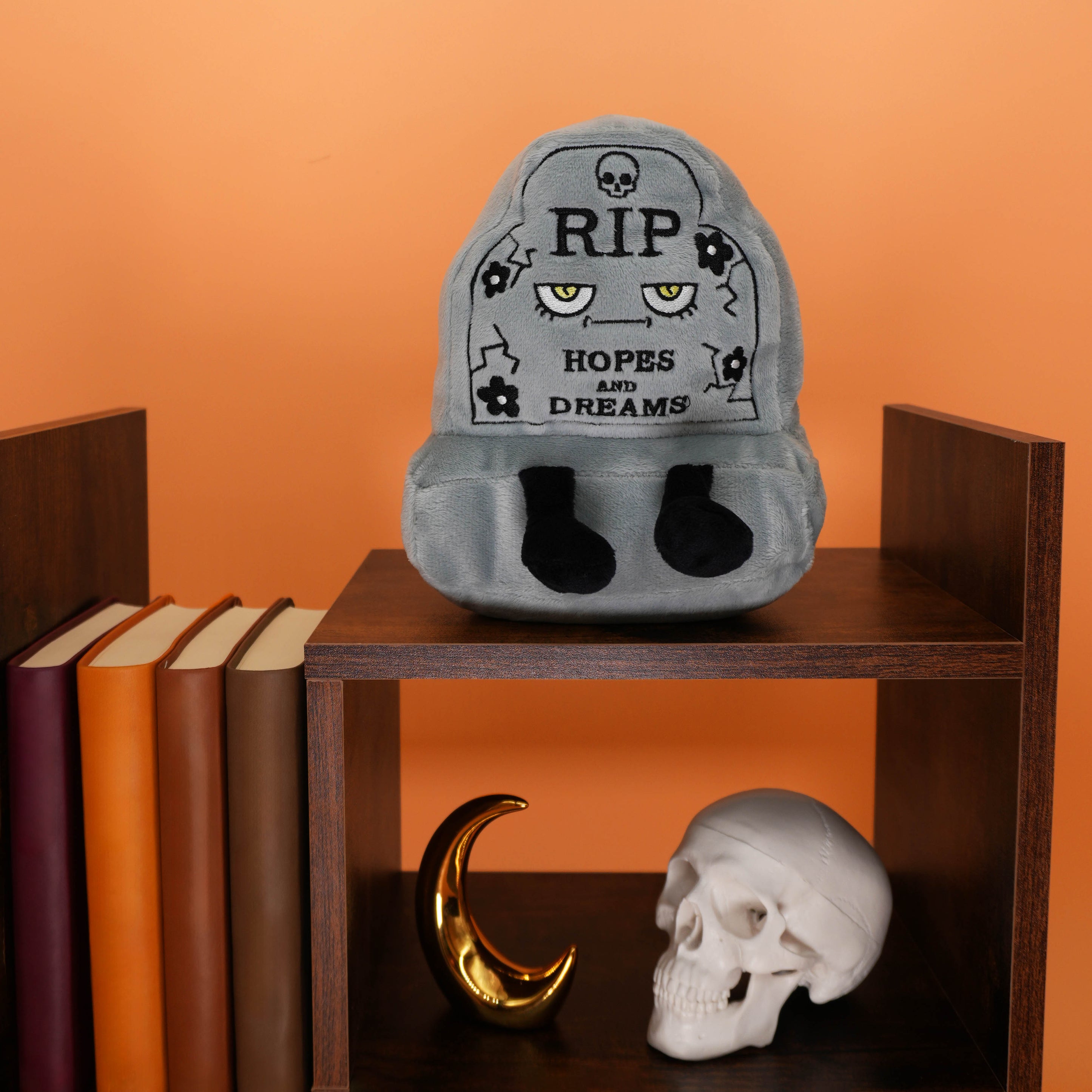 Gray headstone plush toy with text on a wooden shelf against an orange background.