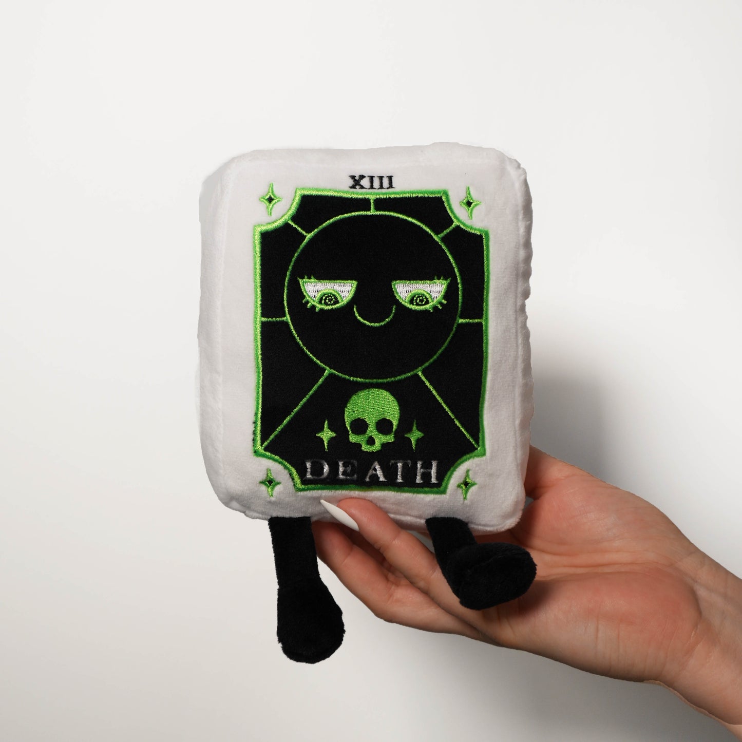 Plush toy with 'Death' tarot card design held by a hand on a white background
