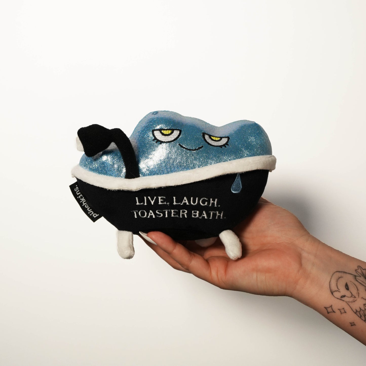 Plush toy resembling a toaster in a bathtub with text 'Live. Laugh. Toaster Bath' held by a hand on a white background.