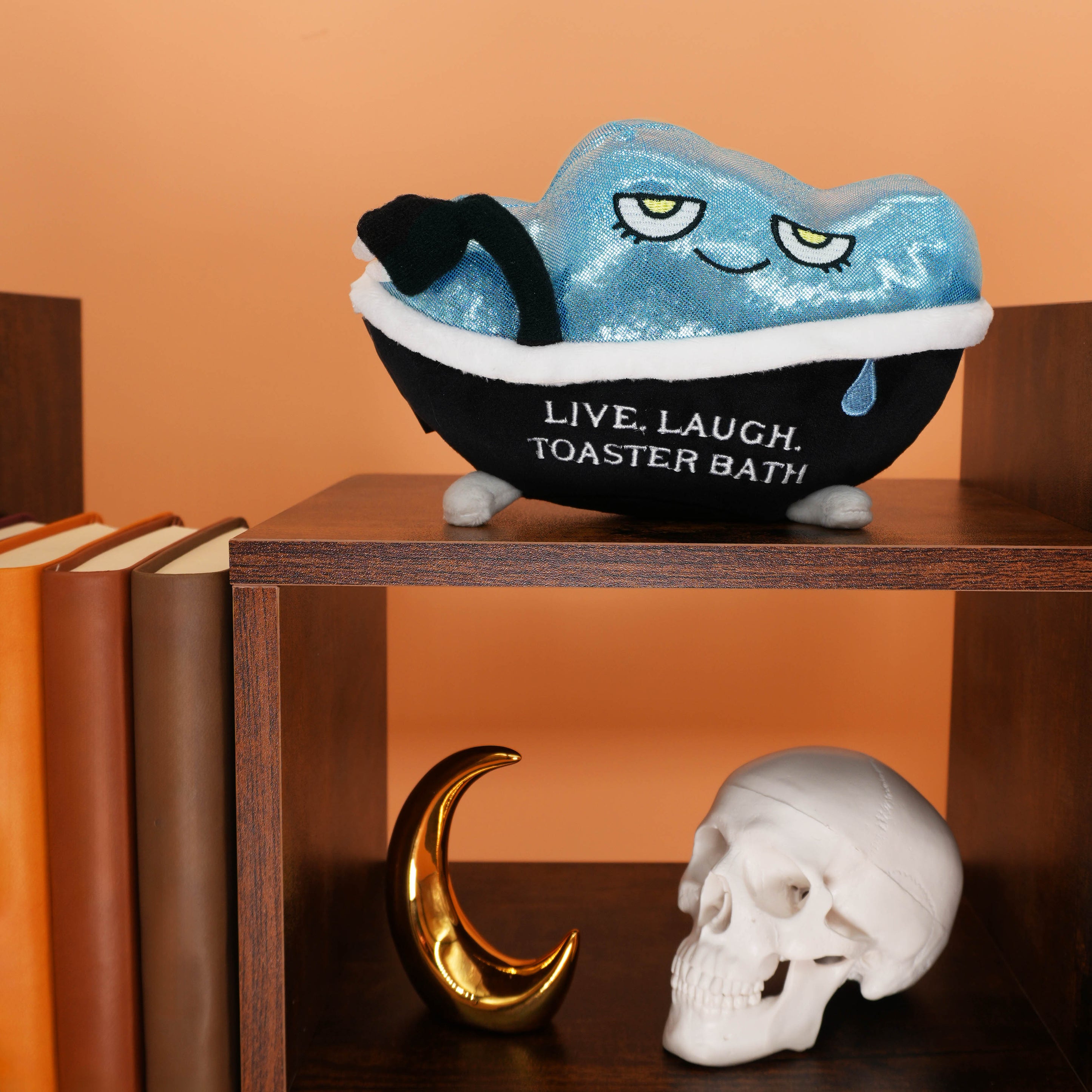 Blue plush toy resembling a toaster  in a bathtub with text on a wooden shelf against an orange wall.