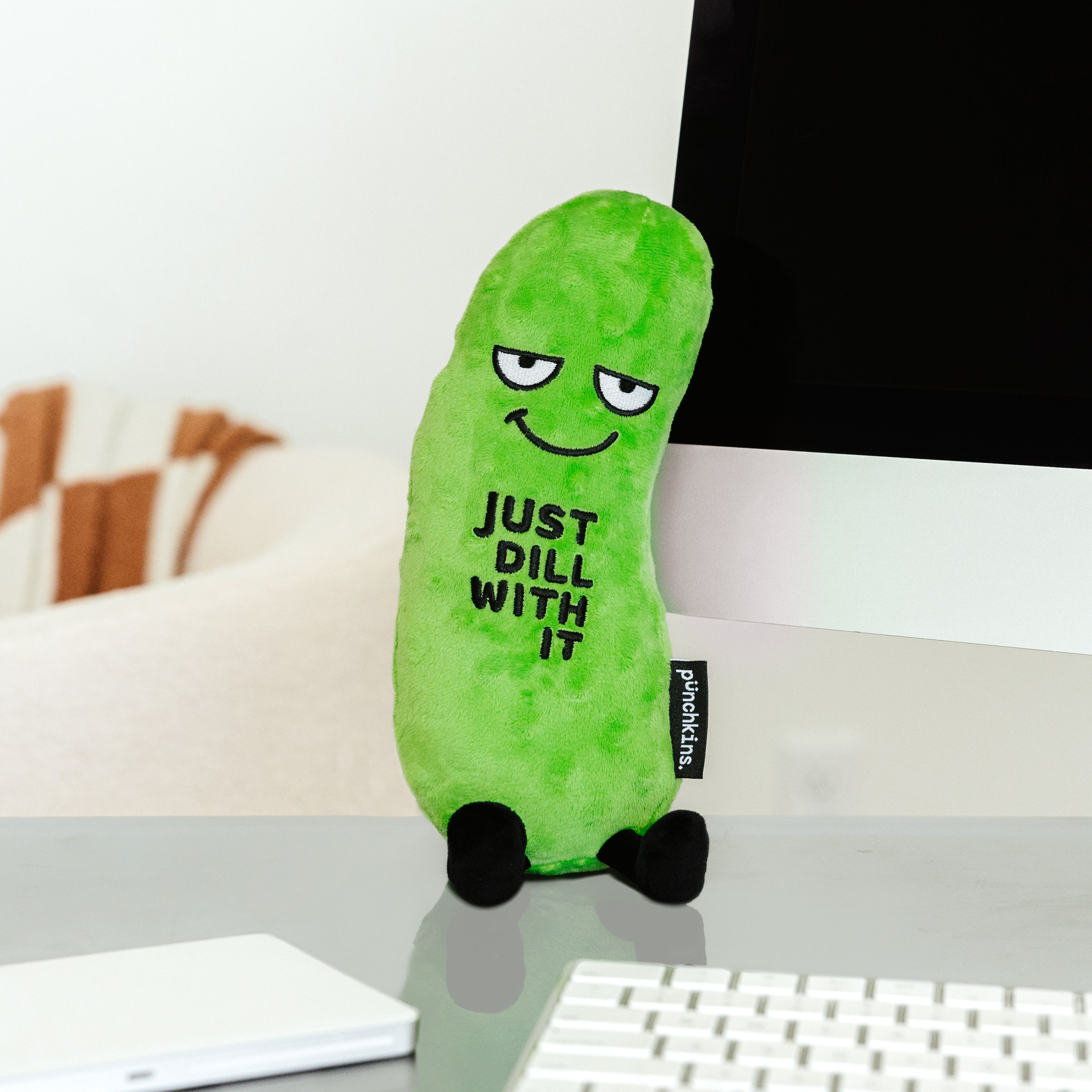 A plush shaped like a pickle with a smiling face and the phrase 'JUST DILL WITH IT' printed on its surface placed on a desk near a computer.