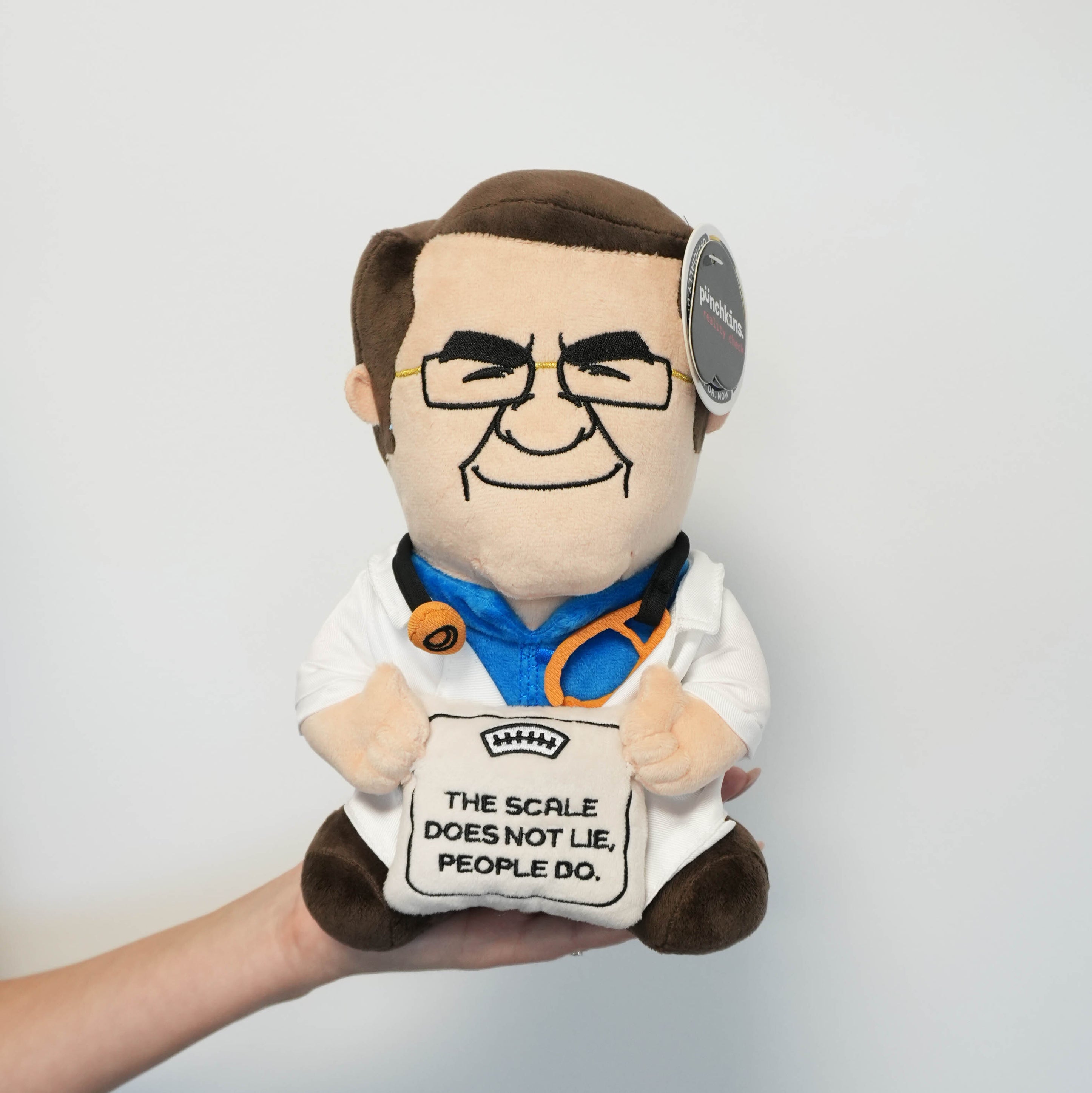 A hand holding a plush toy resembling Dr. Now, who is wearing glasses and a stethoscope and is holding a scale that reads, "The scale does not lie, people do."