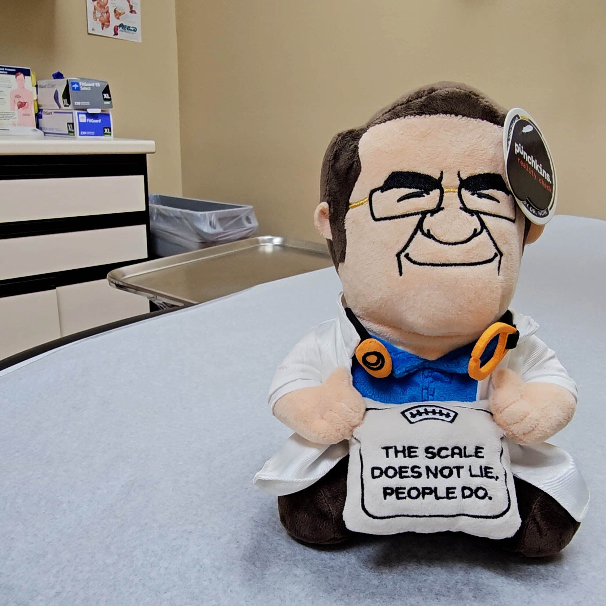 Plush toy resembling Dr. Now, who is wearing glasses and a stethoscope, holding a scale that reads, "The scale does not lie, people do."