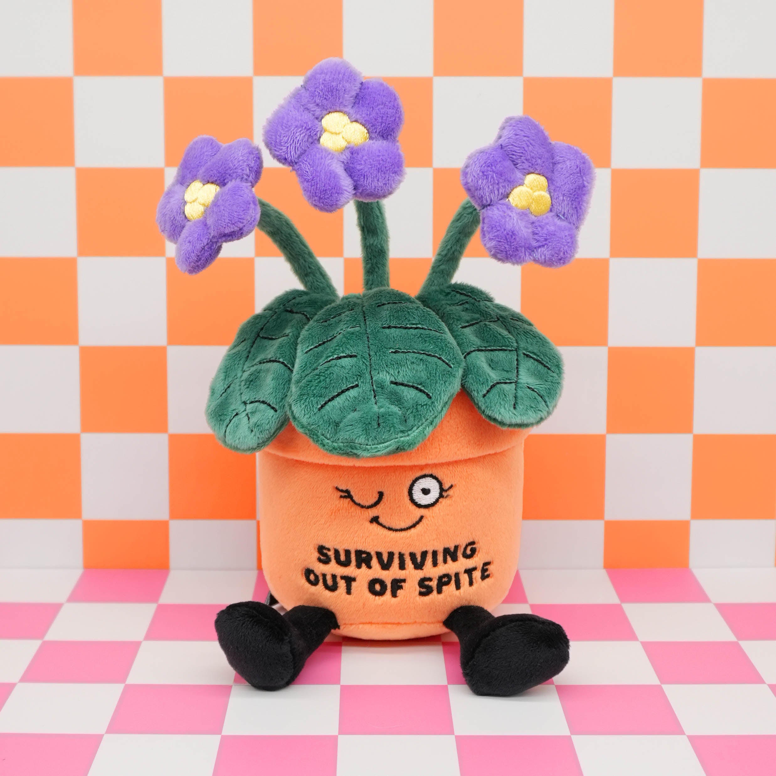 A plush pot of flowers with the text "surviving out of spite" on a checkered background. 