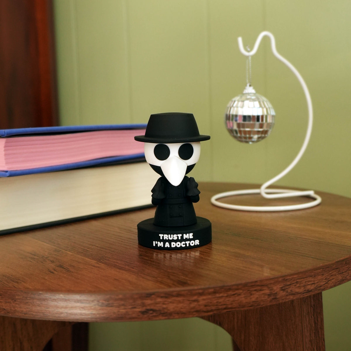 A small black-and-white doctor-themed figurine standing on a round wooden table, with the text 'TRUST ME I'M A DOCTOR' on its base. A book, a white wire stand, and a mirrored ball are visible in the background against an olive-green wall.