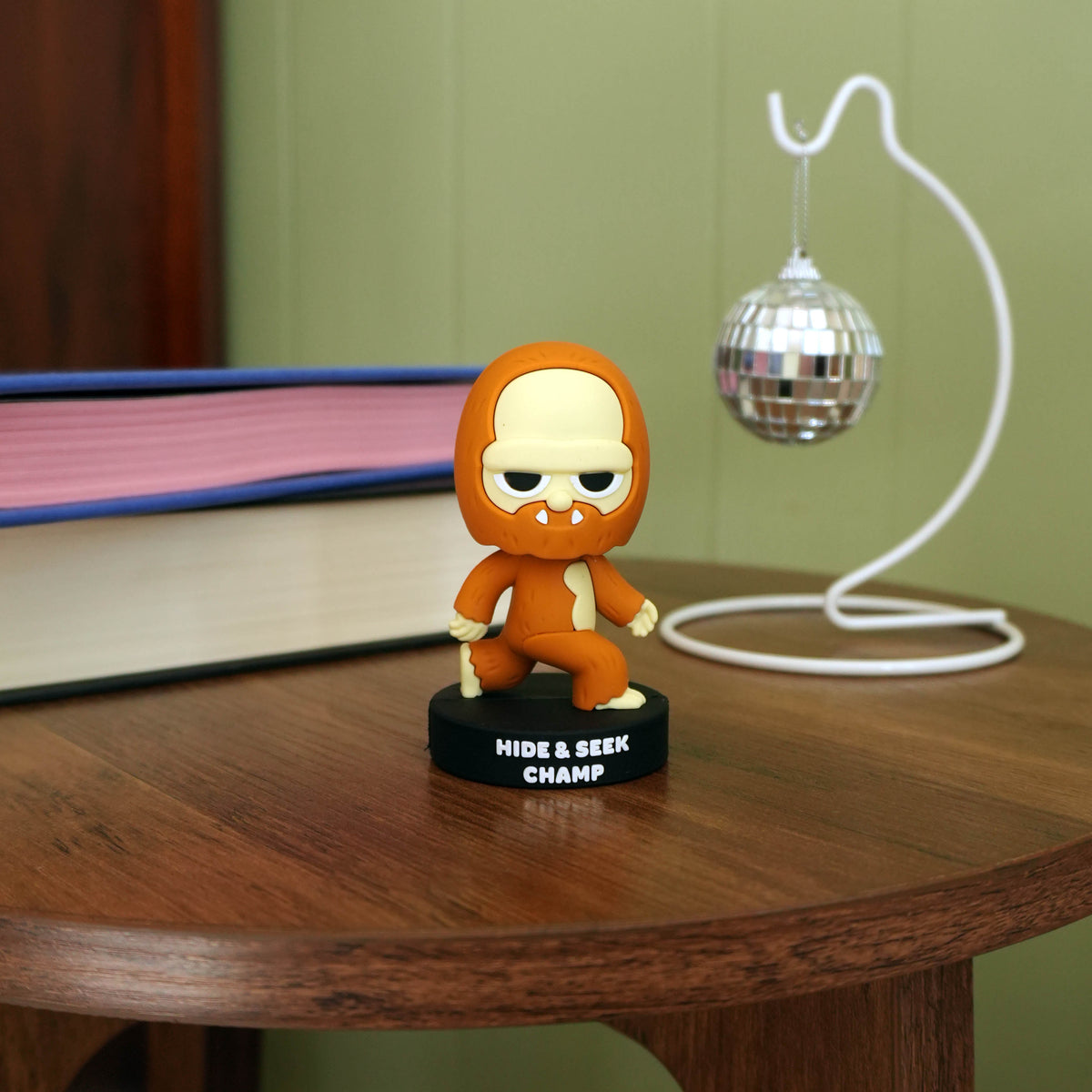 A brown and cream Bigfoot 'HIDE & SEEK CHAMP' bobblehead figure standing on a black base on a wooden table, with a disco ball prop and books in the background.