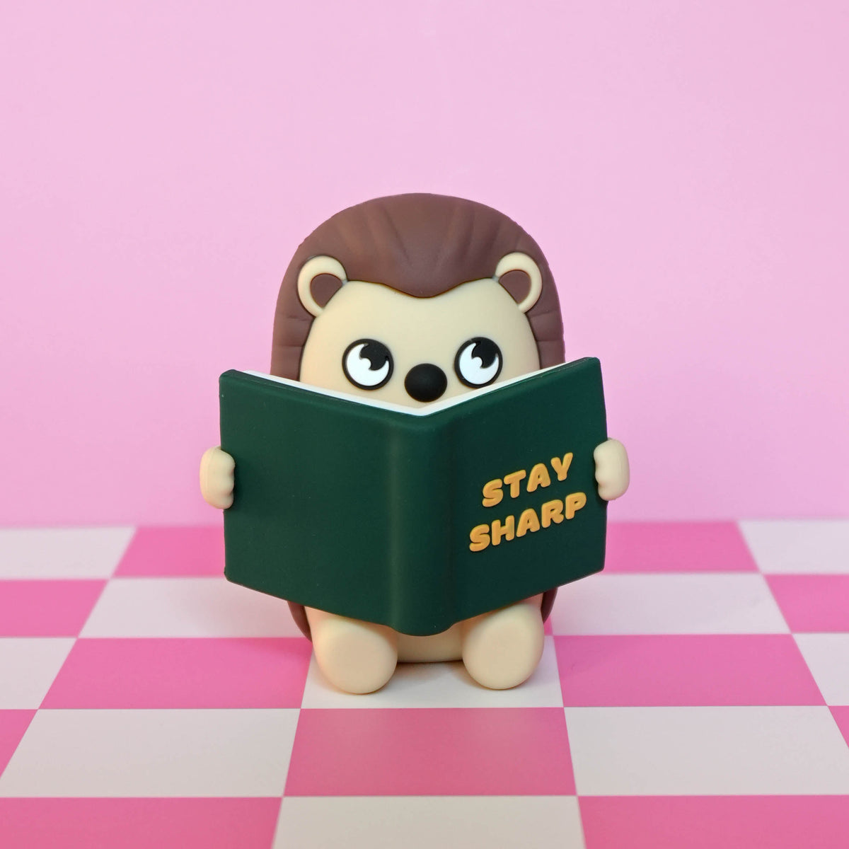A small hedgehog figurine with brown spines and a cream face, holding a dark green book with the words 'STAY SHARP' in yellow. The figurine is centered against a pink background with a pink-and-white checkered floor.
