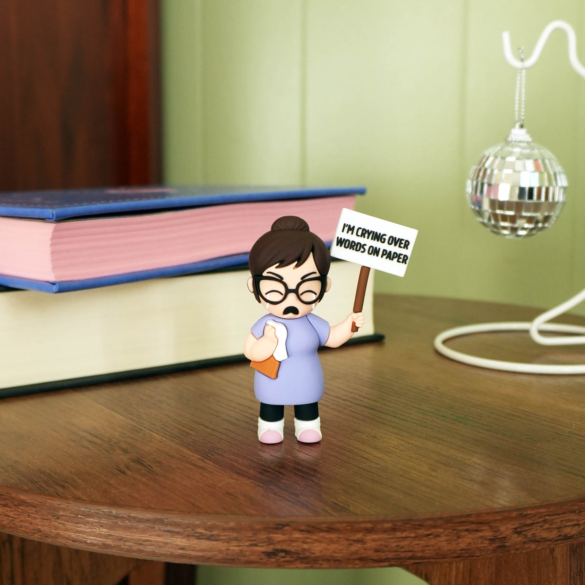 A small figurine of a crying woman holding a sign that reads 'I'M CRYING OVER WORDS ON PAPER' stands on a wooden table next to stacked books, with a disco ball lamp in the background.