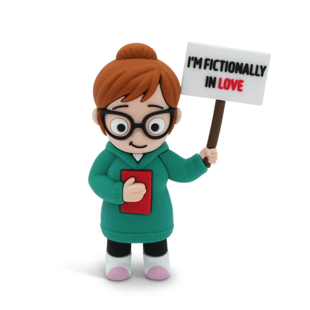 A small figurine of a woman with brown hair in a bun, wearing black glasses and a green sweater, holding a red book and a sign that reads 'I'M FICTIONALLY IN LOVE' on a white background.