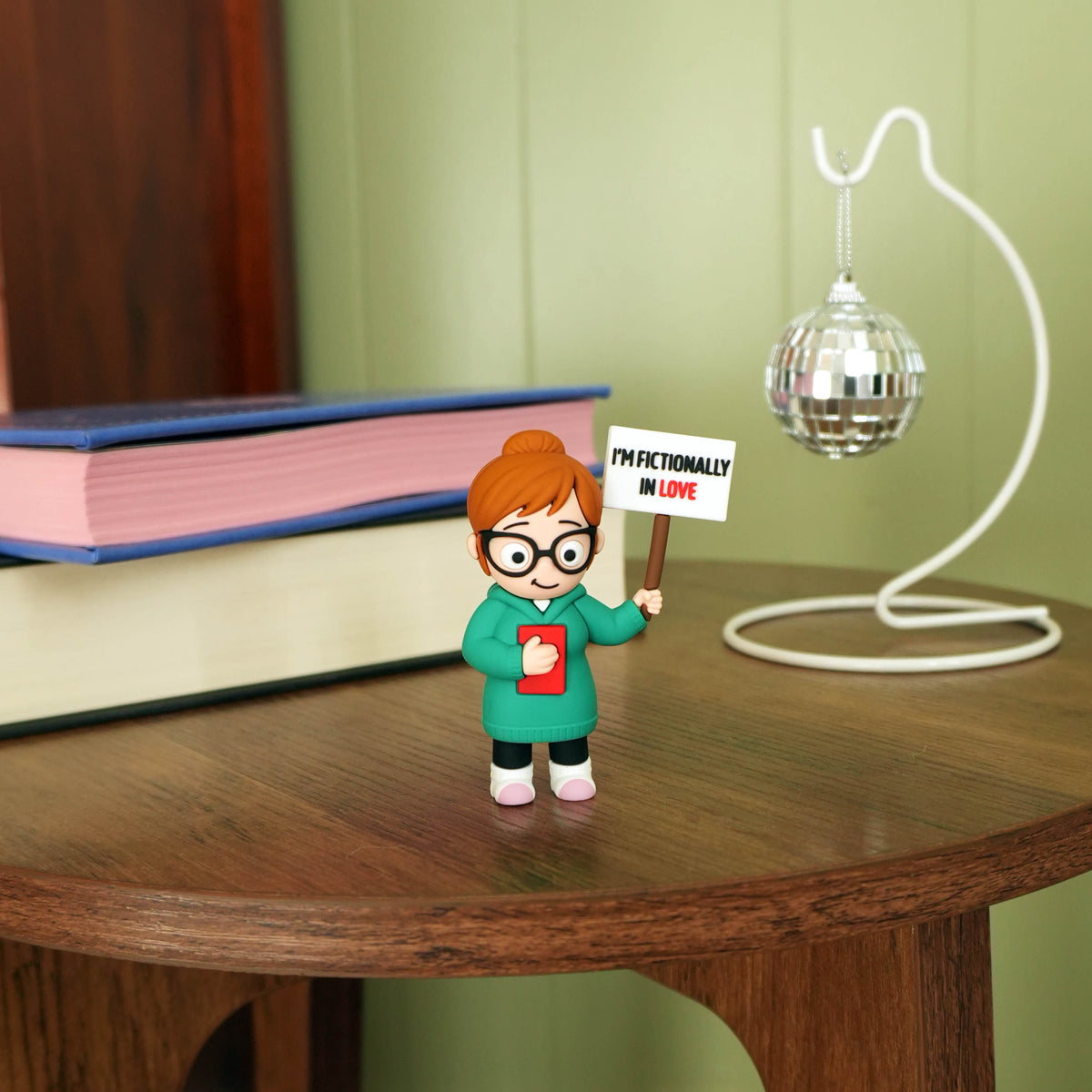 A small figurine of a person with brown hair, glasses, and a green sweater holding a sign that reads 'I'M FICTIONALLY IN LOVE' stands on a wooden table. The background features a green paneled wall, a white disco ball on a stand, and a stack of books.