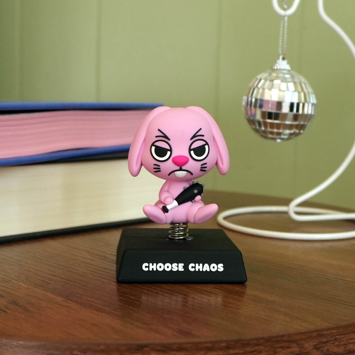 A pink cartoon rabbit figurine with an angry expression, holding a bat with spikes, standing on a black base that reads 'CHOSE CHAOS'. The figurine is placed on a wooden table with a green paneled wall, a disco ball decoration, and books in the background.