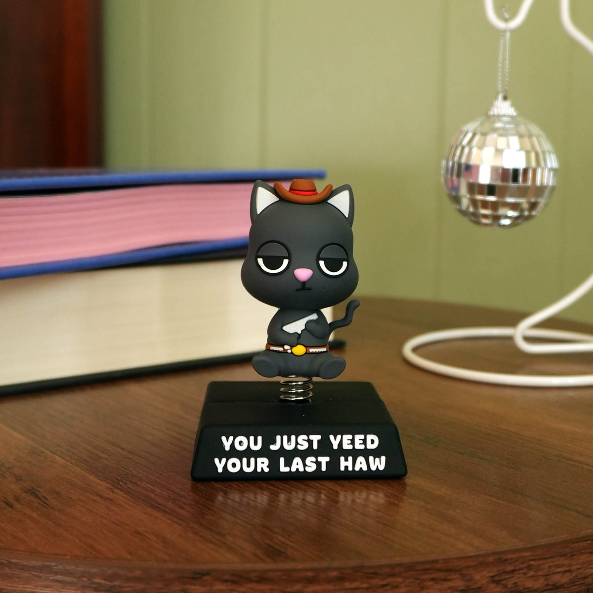 A small cat figurine wearing a brown hat and white shirt, seated on a black base with the text 'YOU JUST YEED YOUR LAST HAW,' placed on a wooden table in a room setting with books and a disco ball in the background.