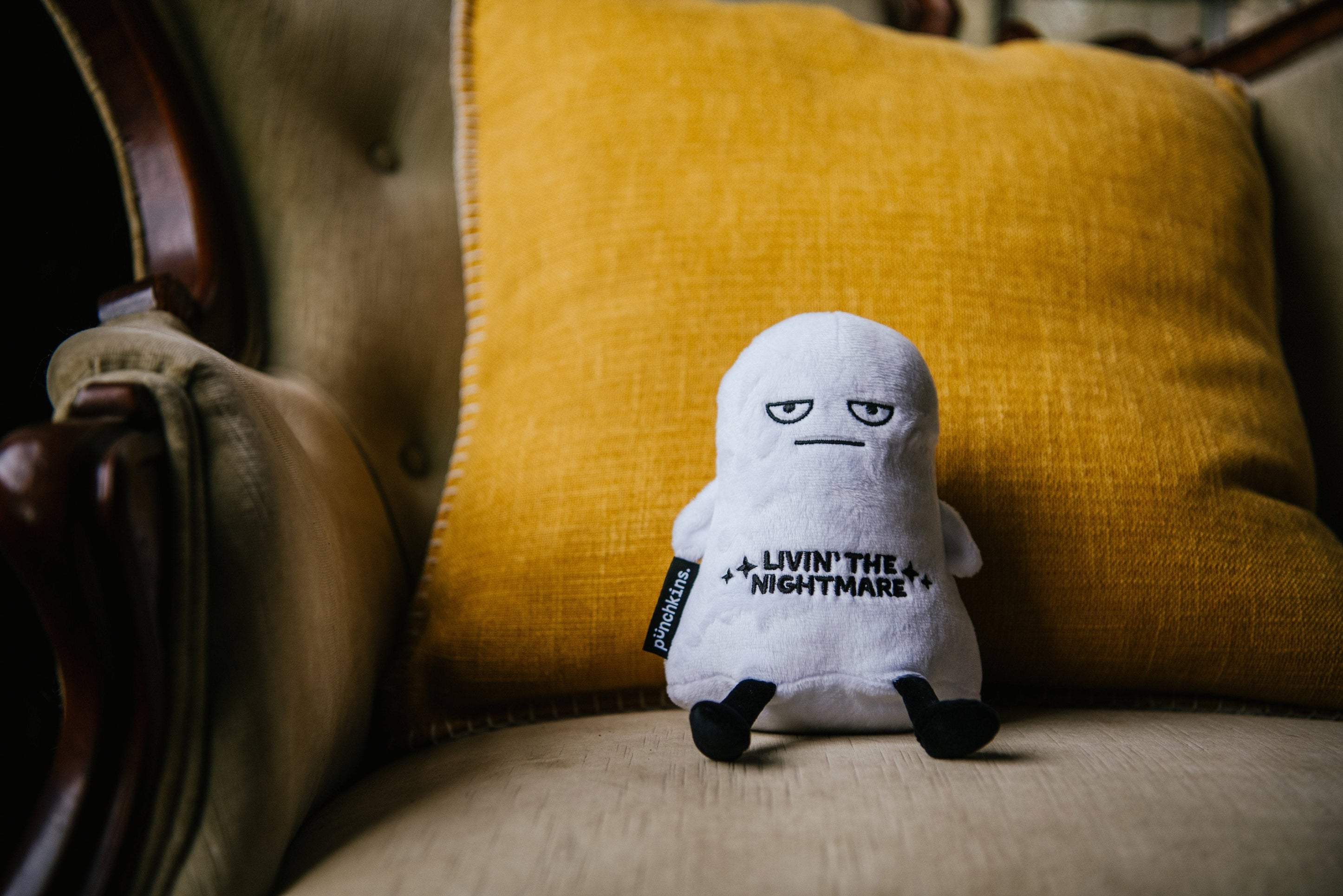 A plush resembling a ghost with a grumpy facial expression and the text 'LIVIN' THE NIGHTMARE' on its chest placed on a couch.