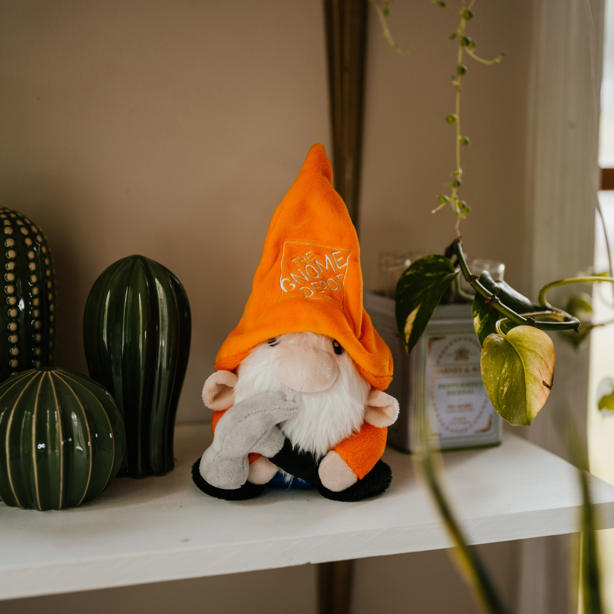 A plush gnome with a white beard wearing an orange pointy hat with 'The Gnome Depot' written on it, sitting on a shelf with a cactus and other decor.