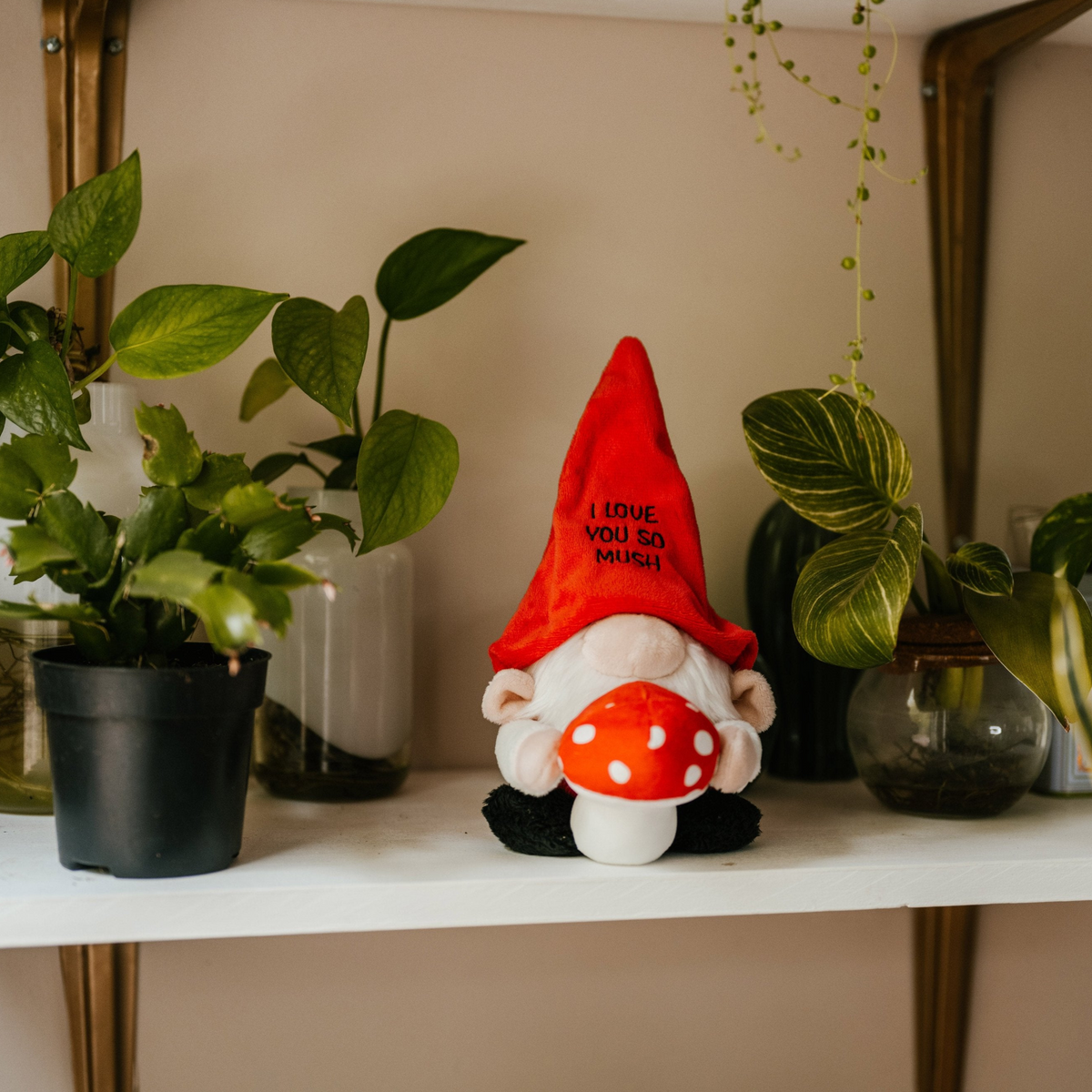 A plush gnome with a red hat with 'I love you so much' text written on it is holding a 3D mushroom. It's placed on a shelf with plants in the background.
