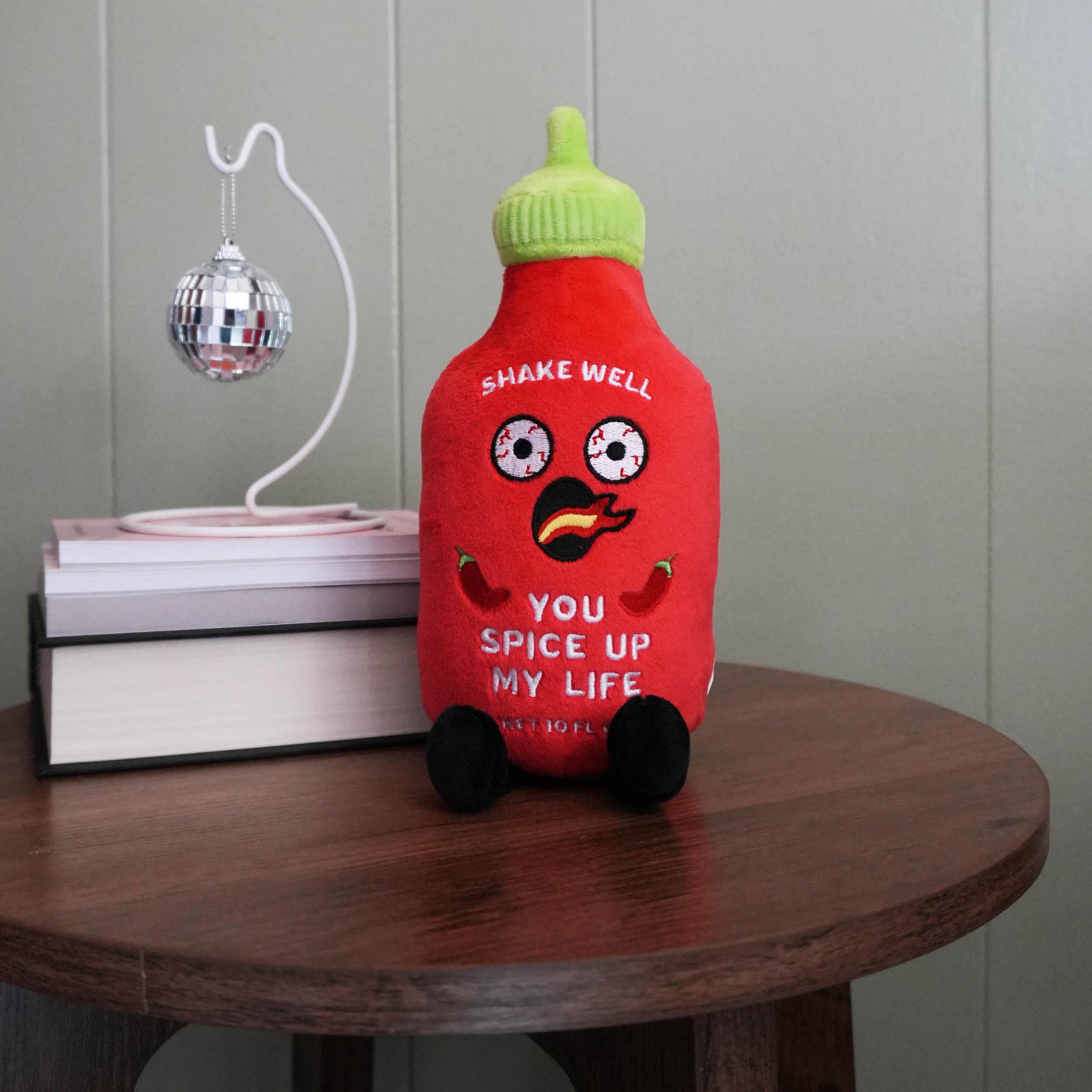 Plush hot sauce bottle with text "YOU SPICE UP MY LIFE" on a table with books and a disco ball. 
