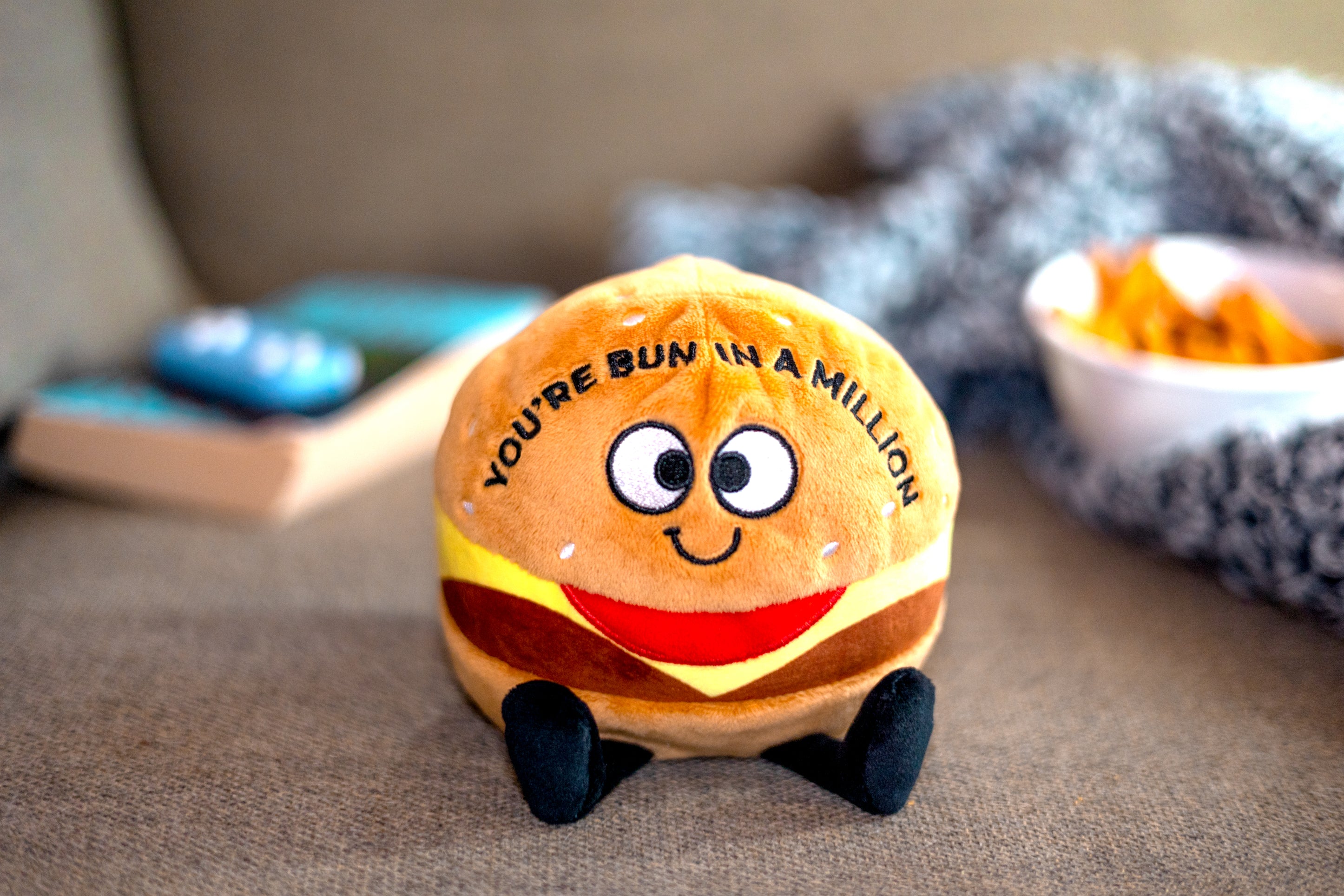 A plush designed to look like a cheeseburger with a smiling face, eyes, and dangly legs. Text on it reads, "You're bun in a million." It's placed on a couch.