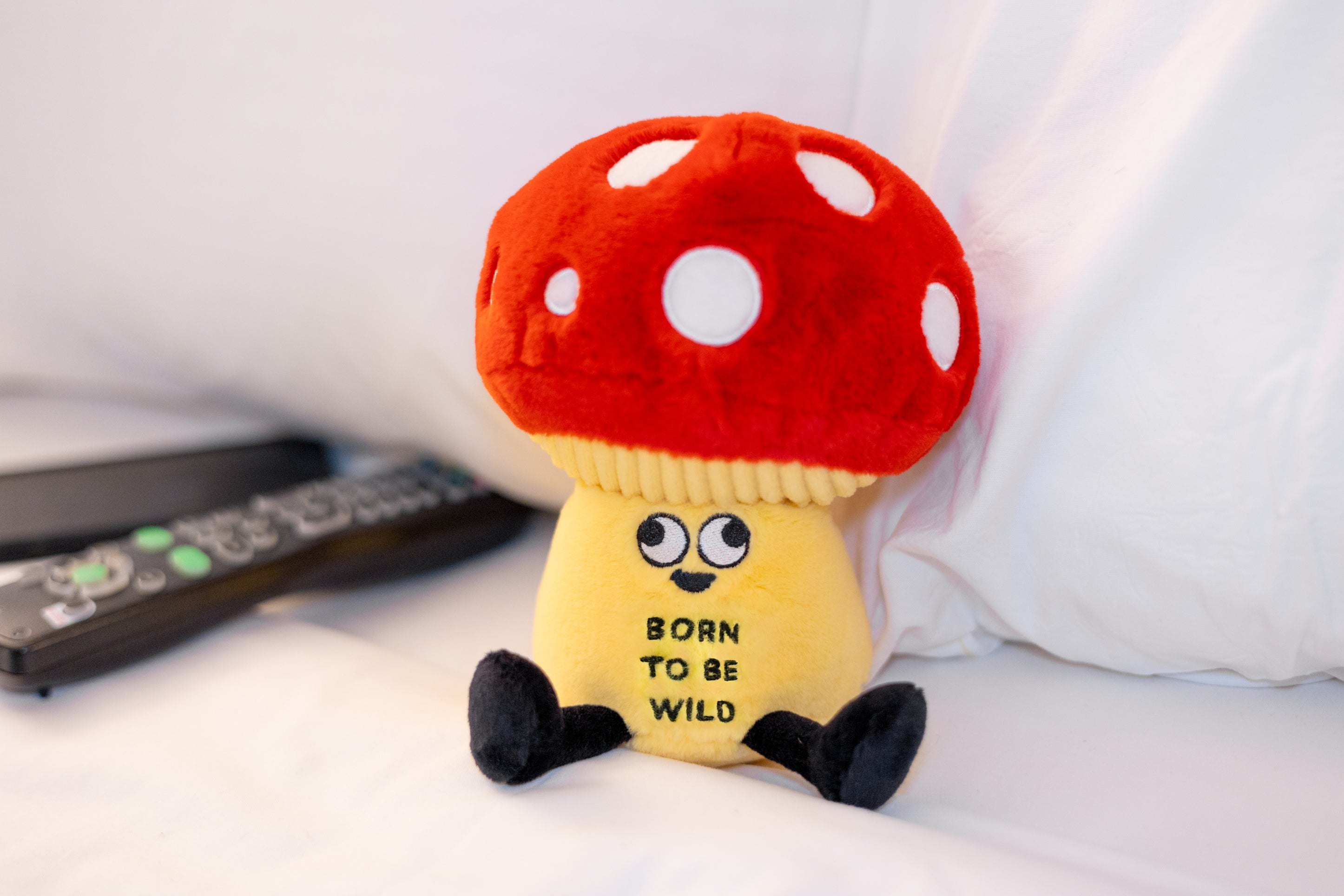 A plush shaped like a mushroom with a face and text on the stem that says 'born to be wild' placed on a bed.