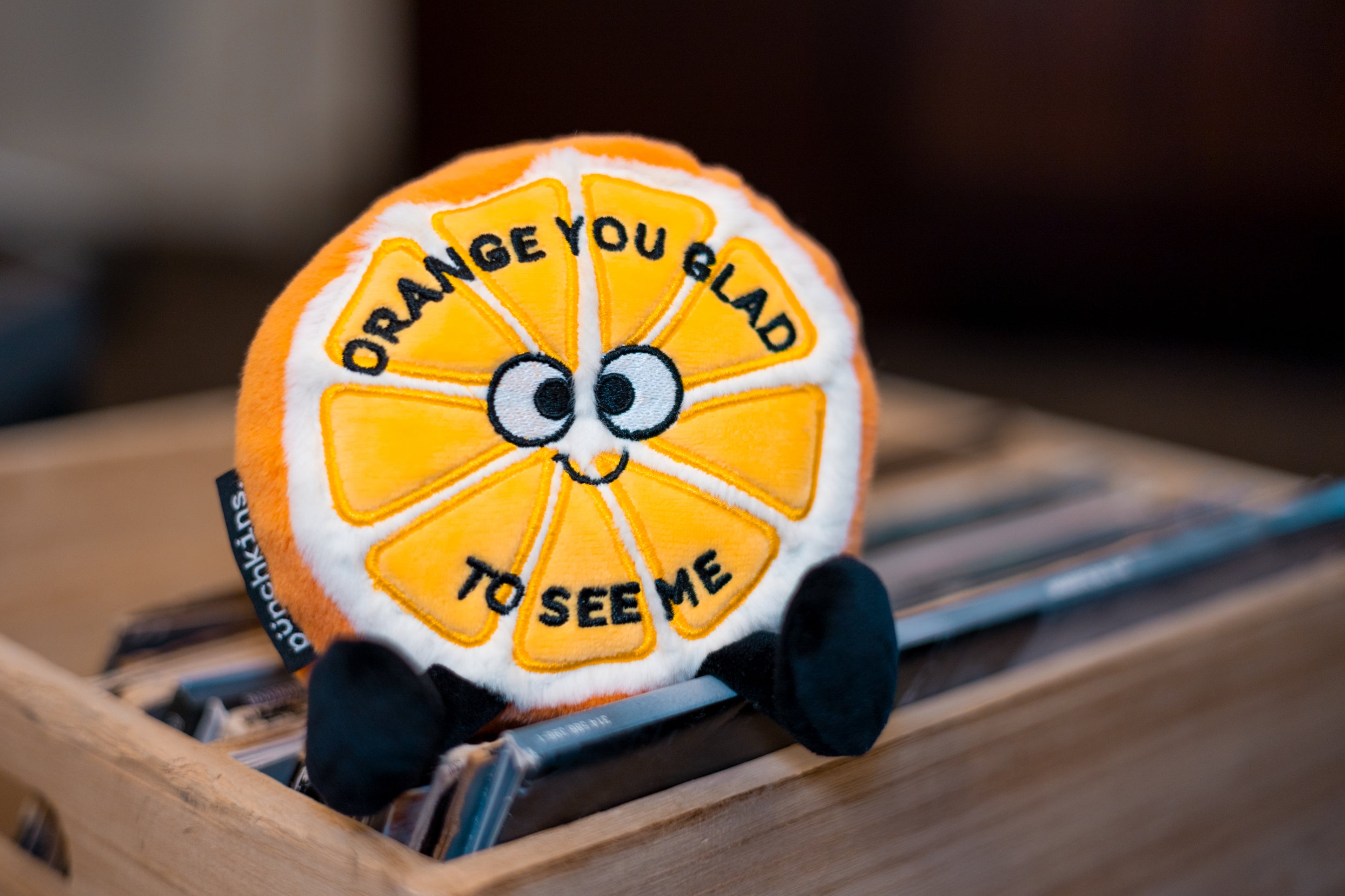 A plush designed to look like an orange with a smiling face and the phrase 'orange you glad to see me' written on it. It's placed on a set of records. 