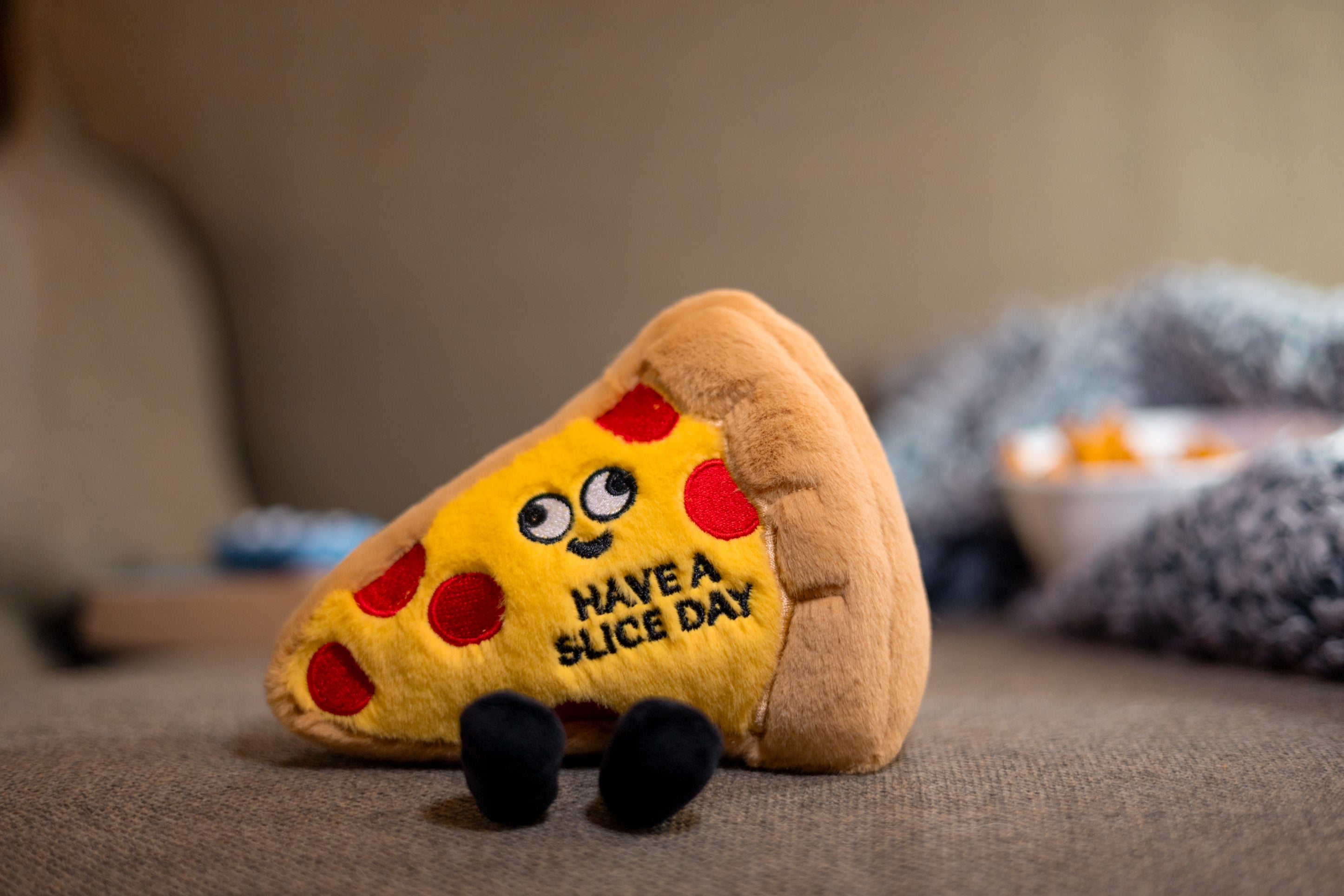 A plush in the shape of a slice of pepperoni pizza with a smiling face and the phrase 'HAVE A SLICE DAY' written on it. It's been placed on a couch.