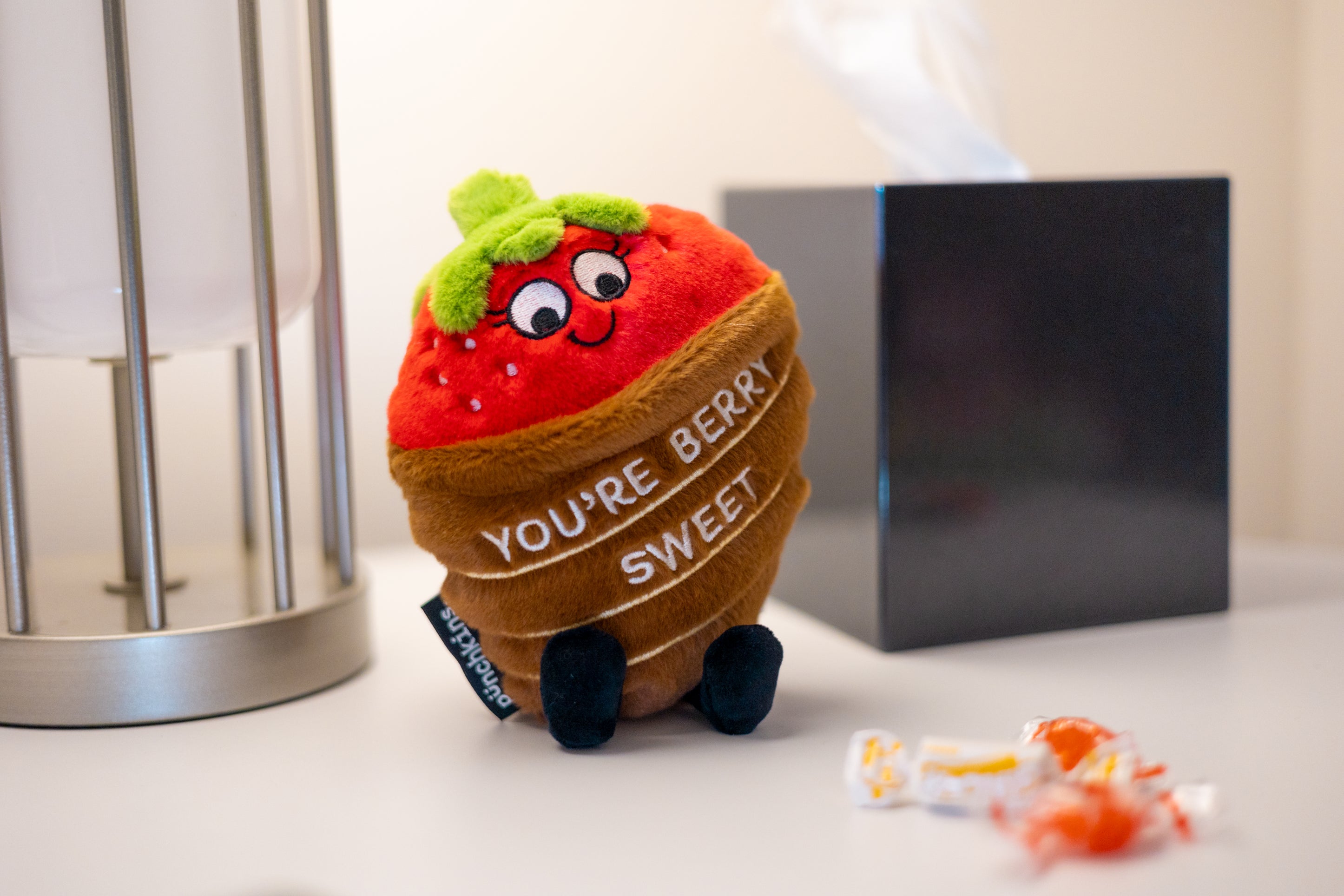 A plush resembling a chocolate covered strawberry placed on a dresser. It has eyes and dangly legs, and there is text on the base that reads 'YOU'RE BERRY SWEET'. 
