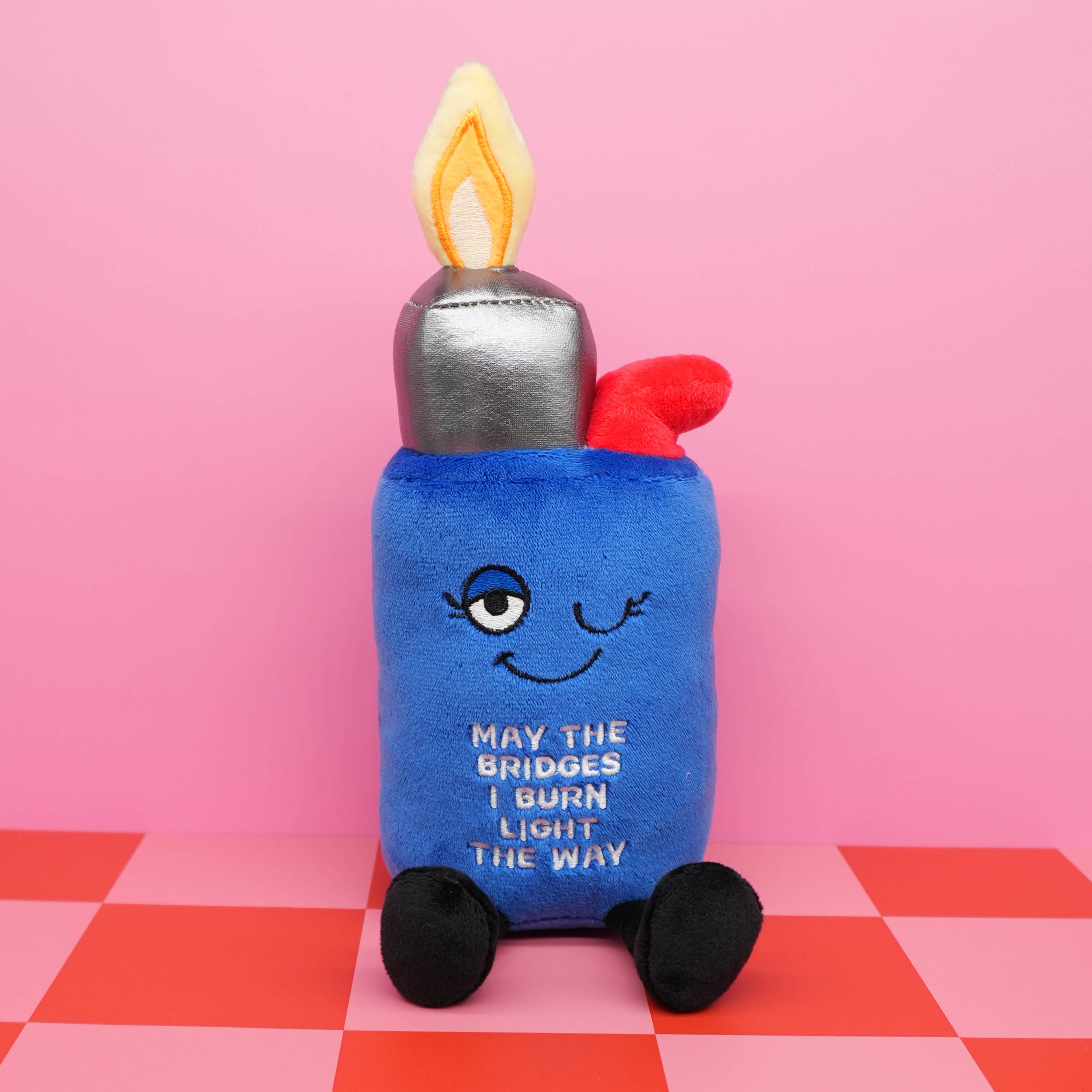 A plush lighter that's on fire and features the text "May the bridges I burn light the way" placed on a checkered background.