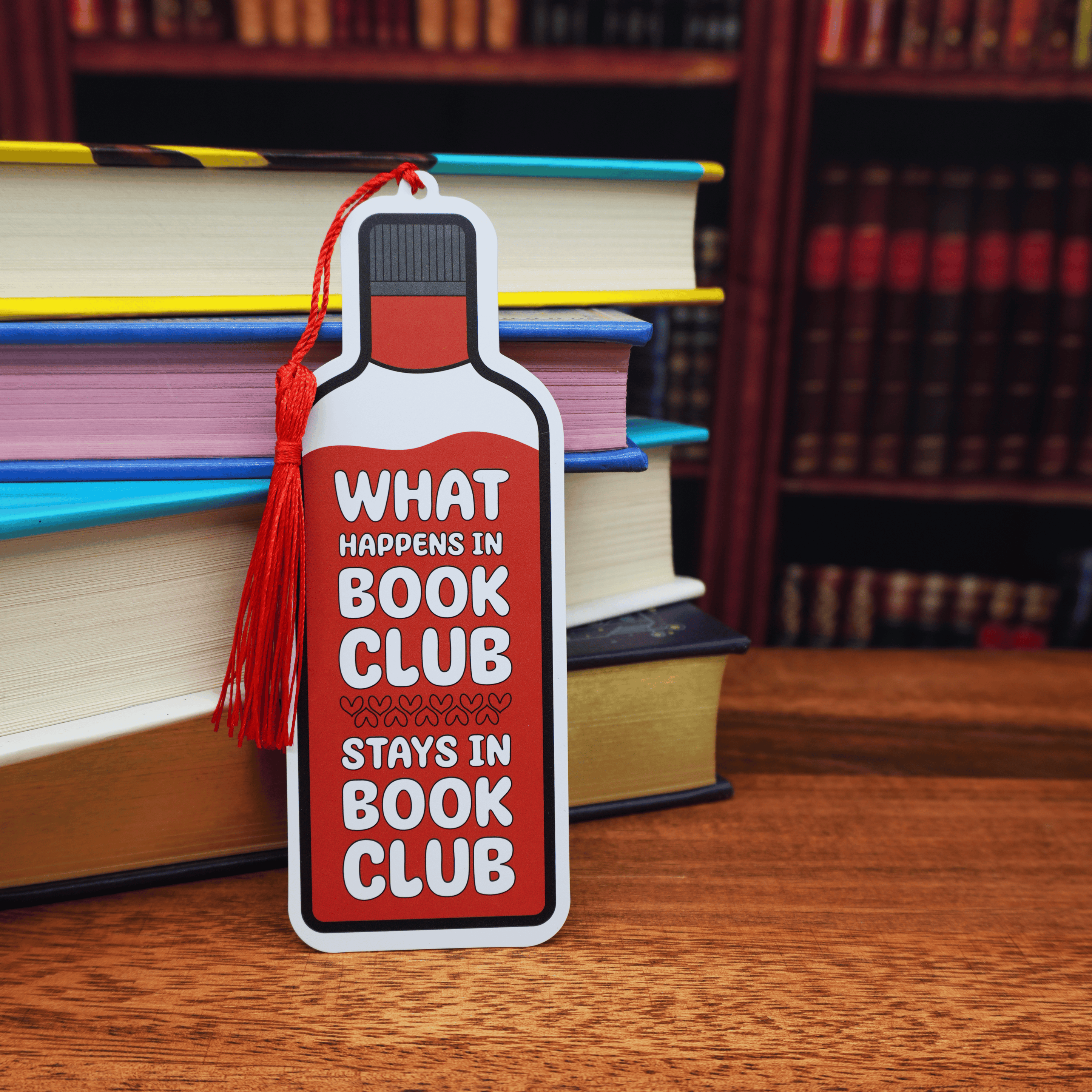 Bookmark shaped like a wine bottle with text 'What happens in book club stays in book club' on a wooden surface with books in the background.