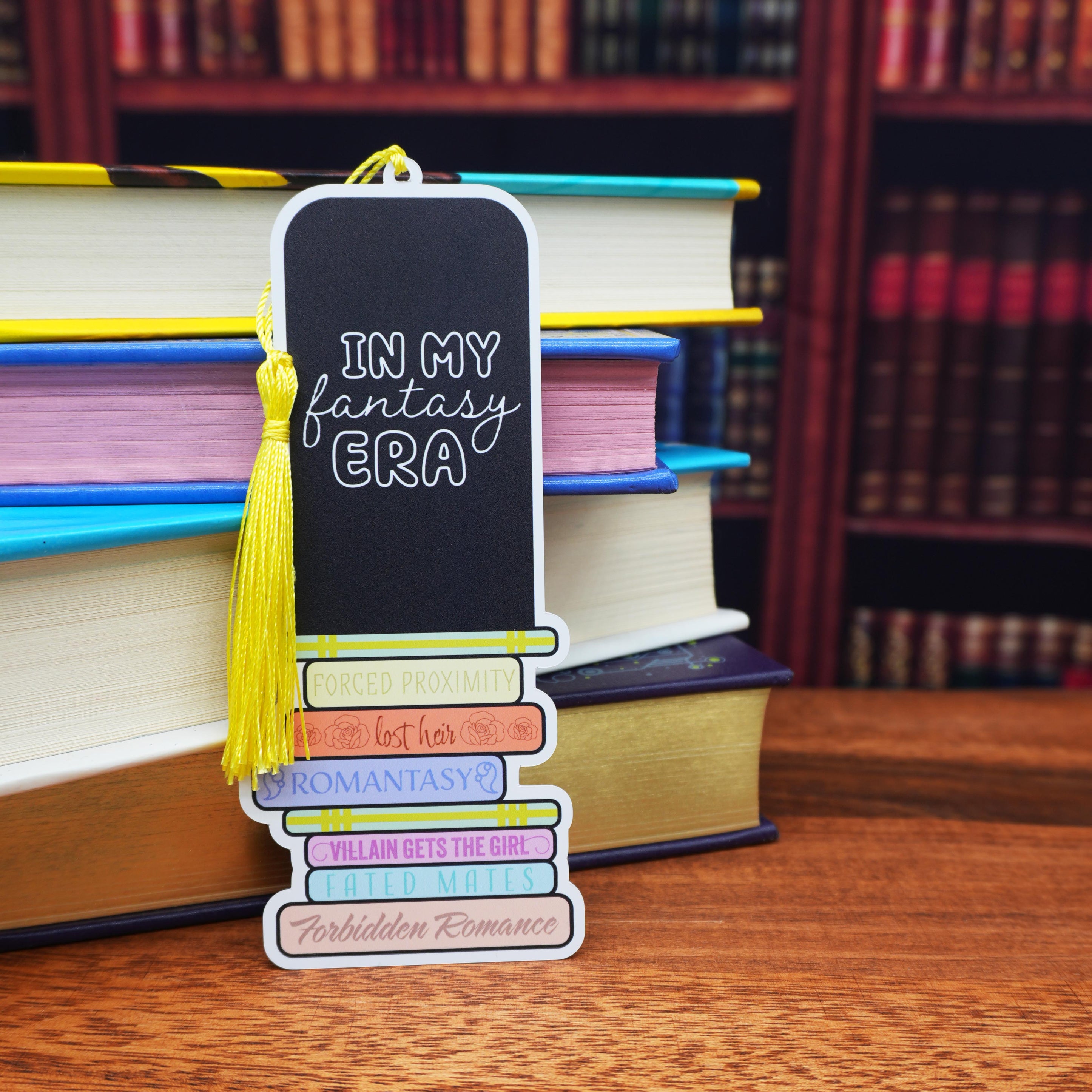 Bookmark with 'In My Fantasy Era' text and tassel on a stack of books with a bookshelf background.