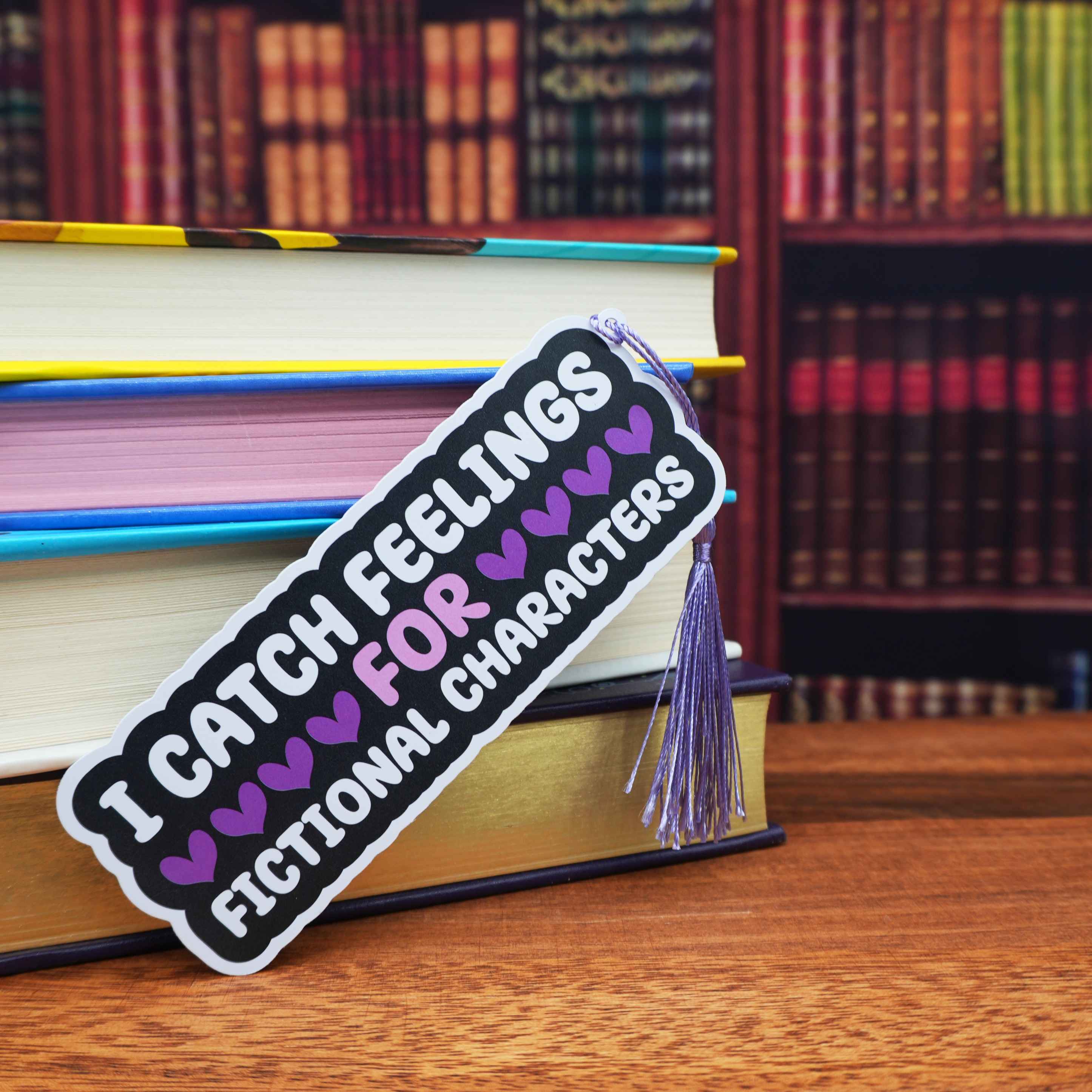 Bookmark with text 'I catch feelings for fictional characters' on a stack of books with a bookshelf background.
