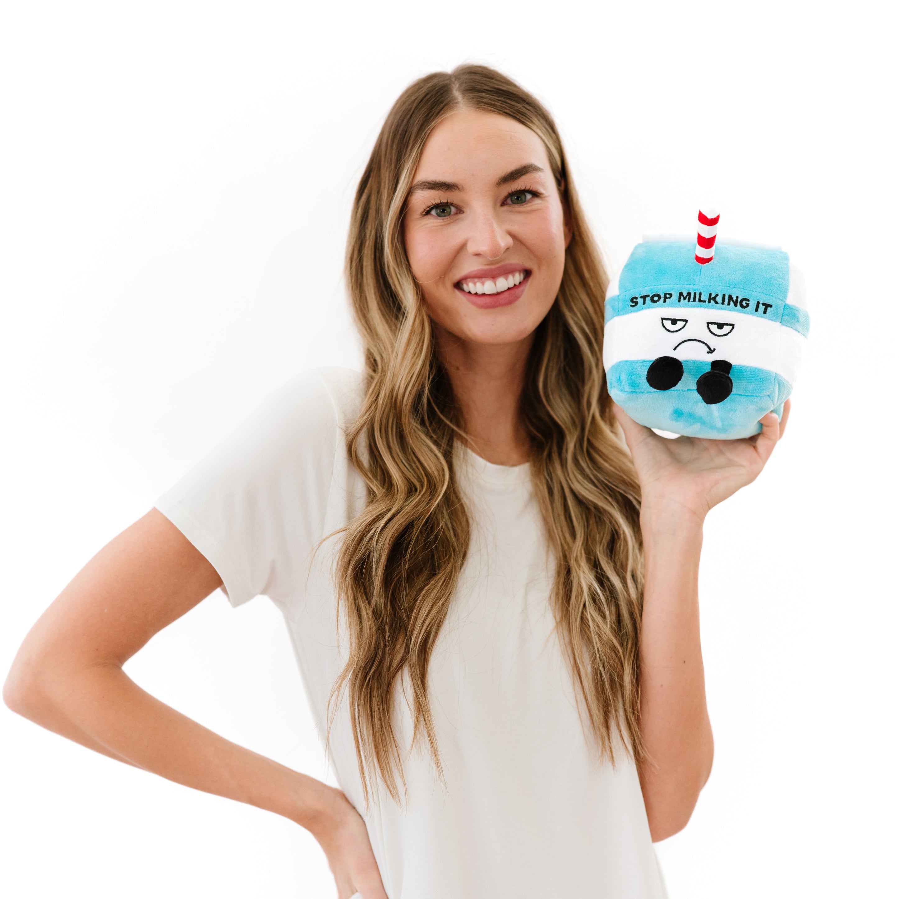 A woman holding a plush designed to resemble a milk carton with a face, dangly legs, and a red and white striped straw. Written on it is the text 'Stop Milking It.'