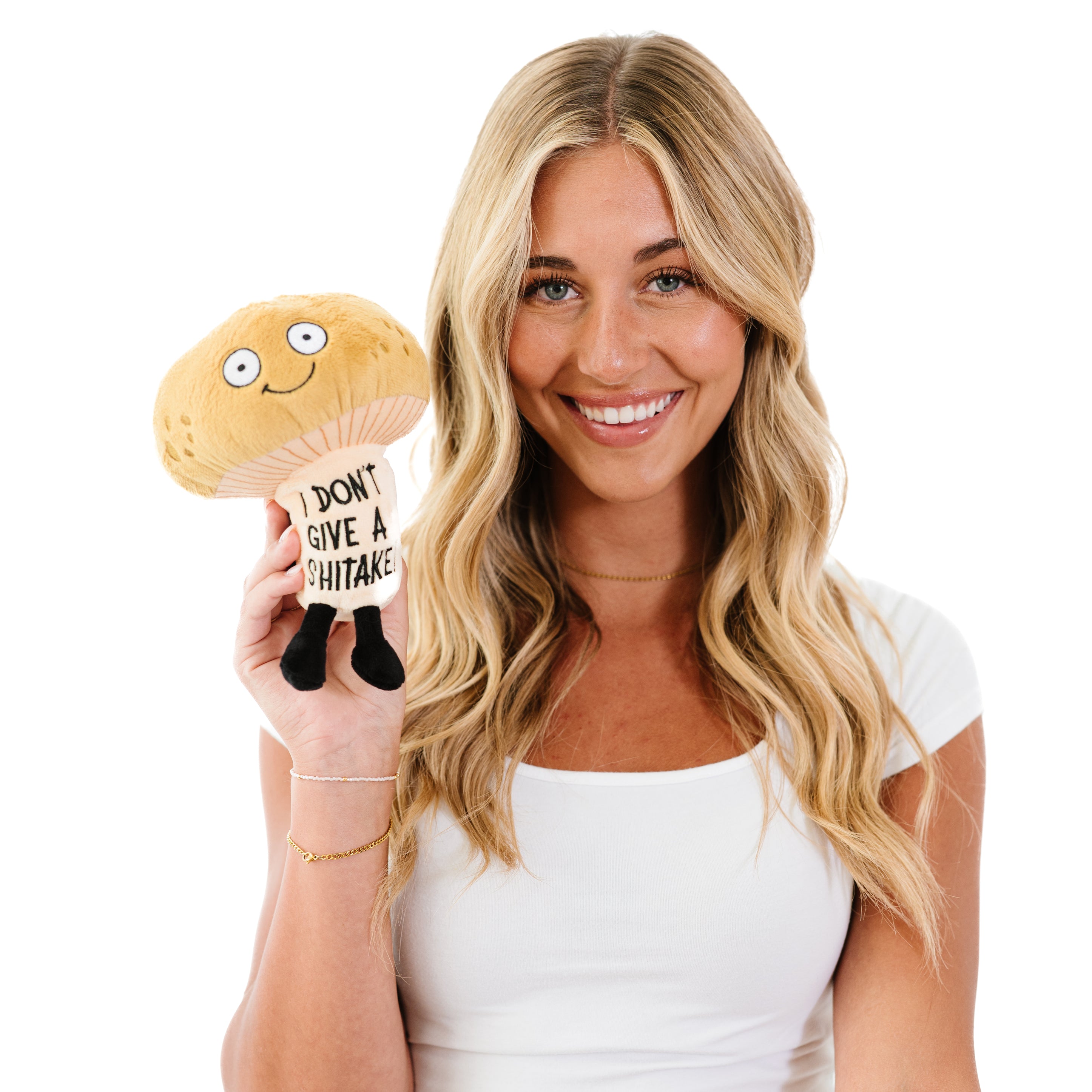 A blonde holding a plush shaped like a mushroom with a smiling face and a text tag that reads 'I DON'T GIVE A SHITAKE'.