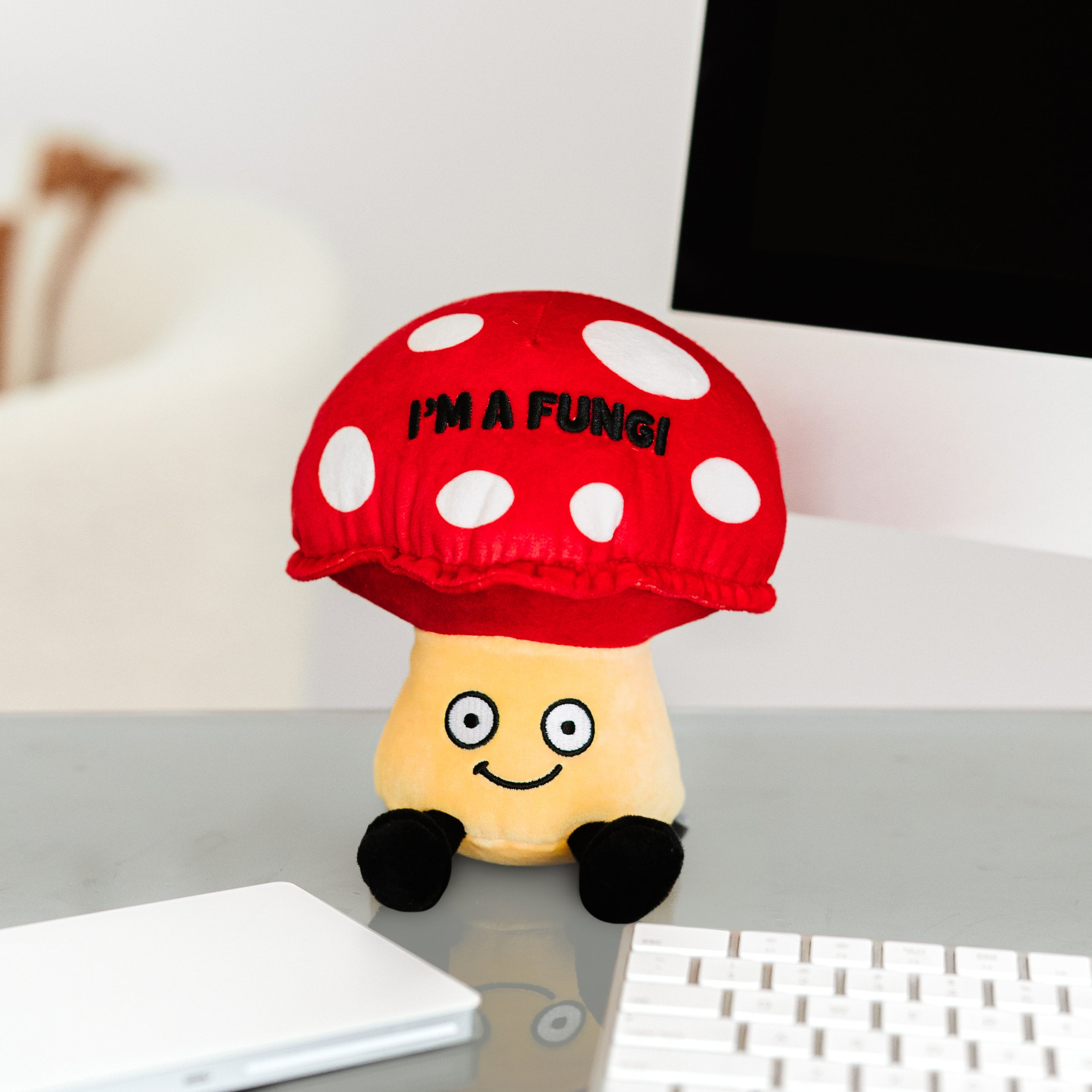 A plush shaped like a red and yellow mushroom with a smiling face and the text 'I'M A FUNGI' on its cap placed on a desk near a computer.