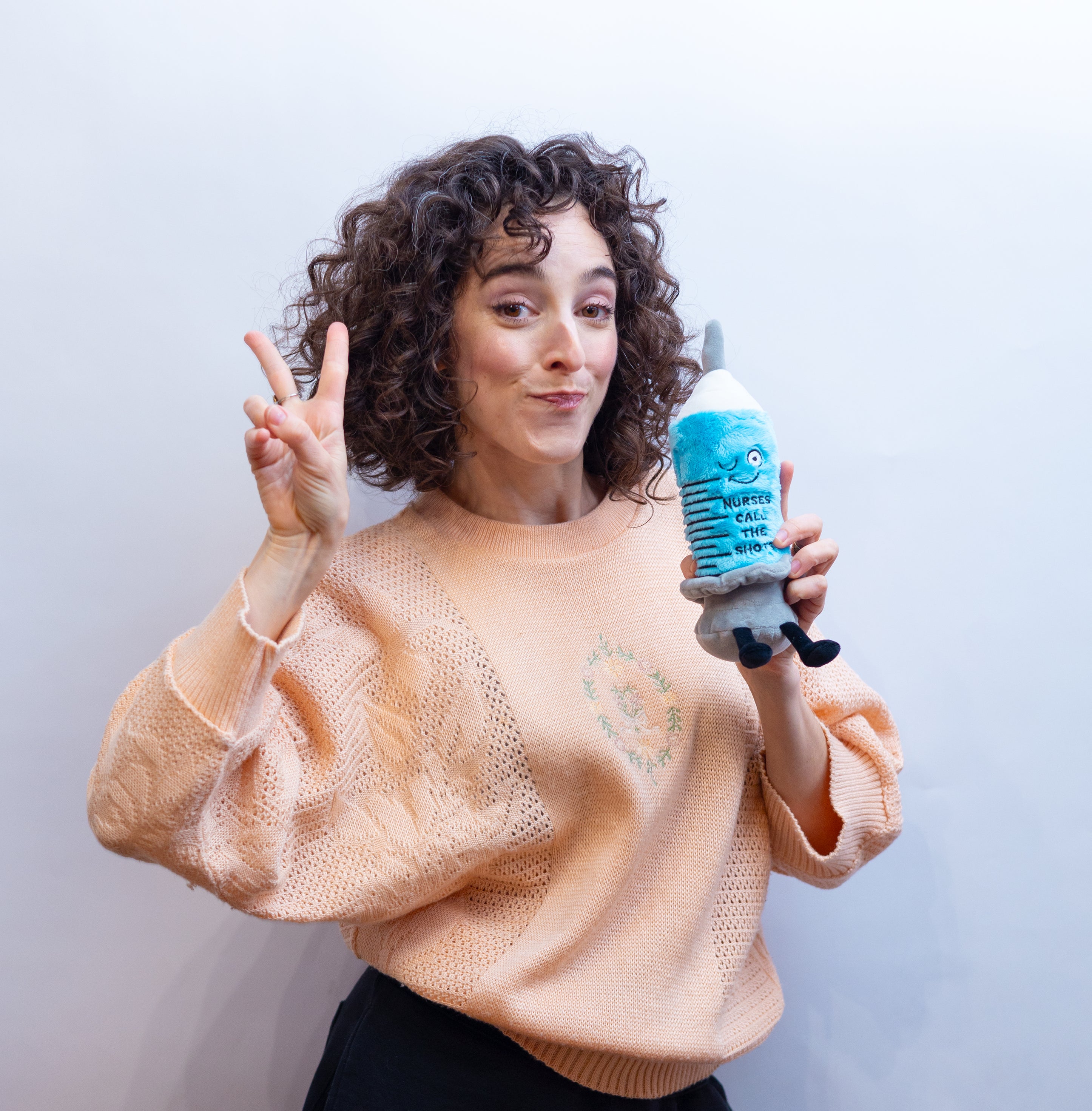 A woman holding a plush designed to resemble a syringe with a smiling face and the text 'Nurses call the shot' on it.