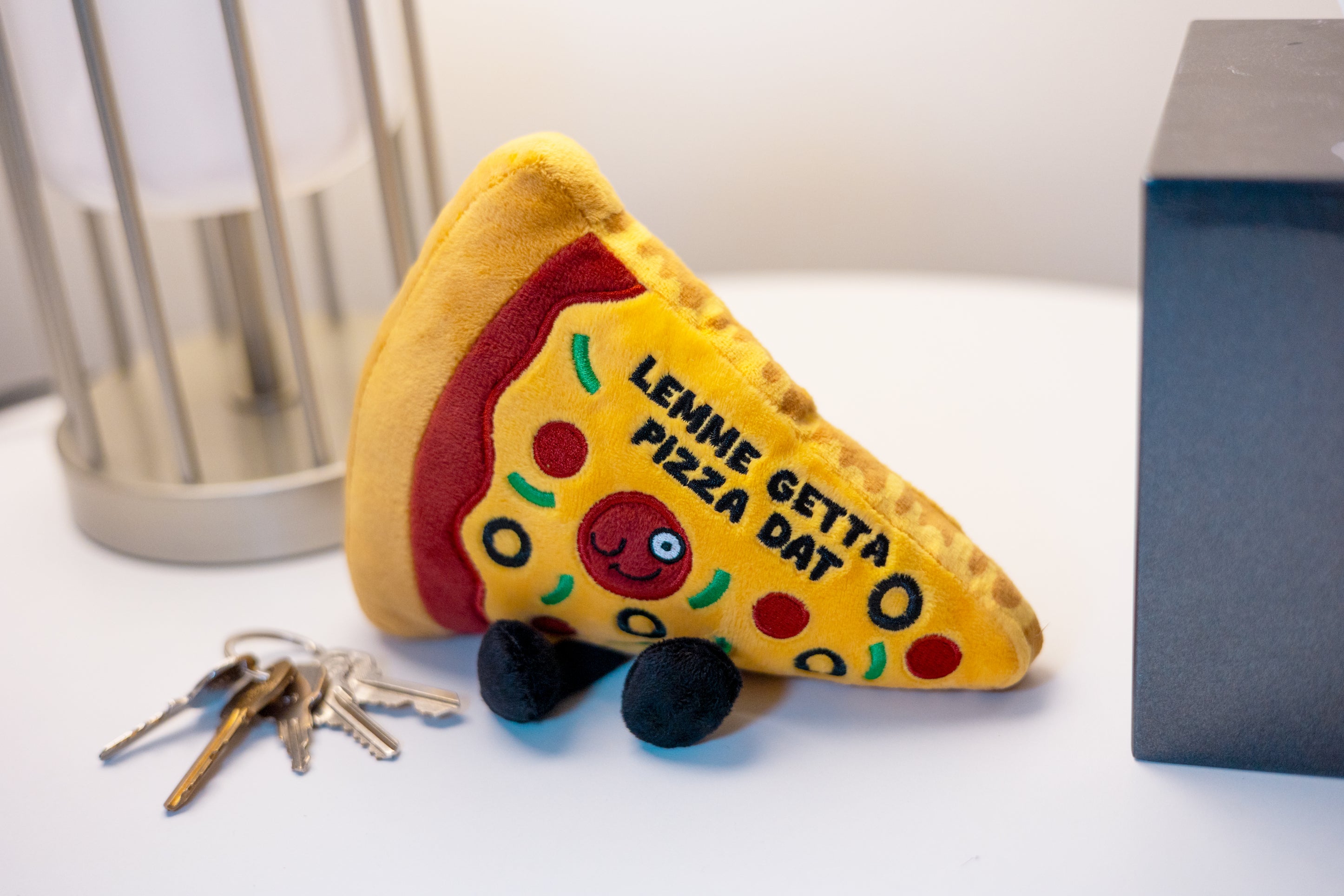 A plush shaped like a slice of pizza with the text 'Lemme get a pizza dat' written on it placed on a desk.