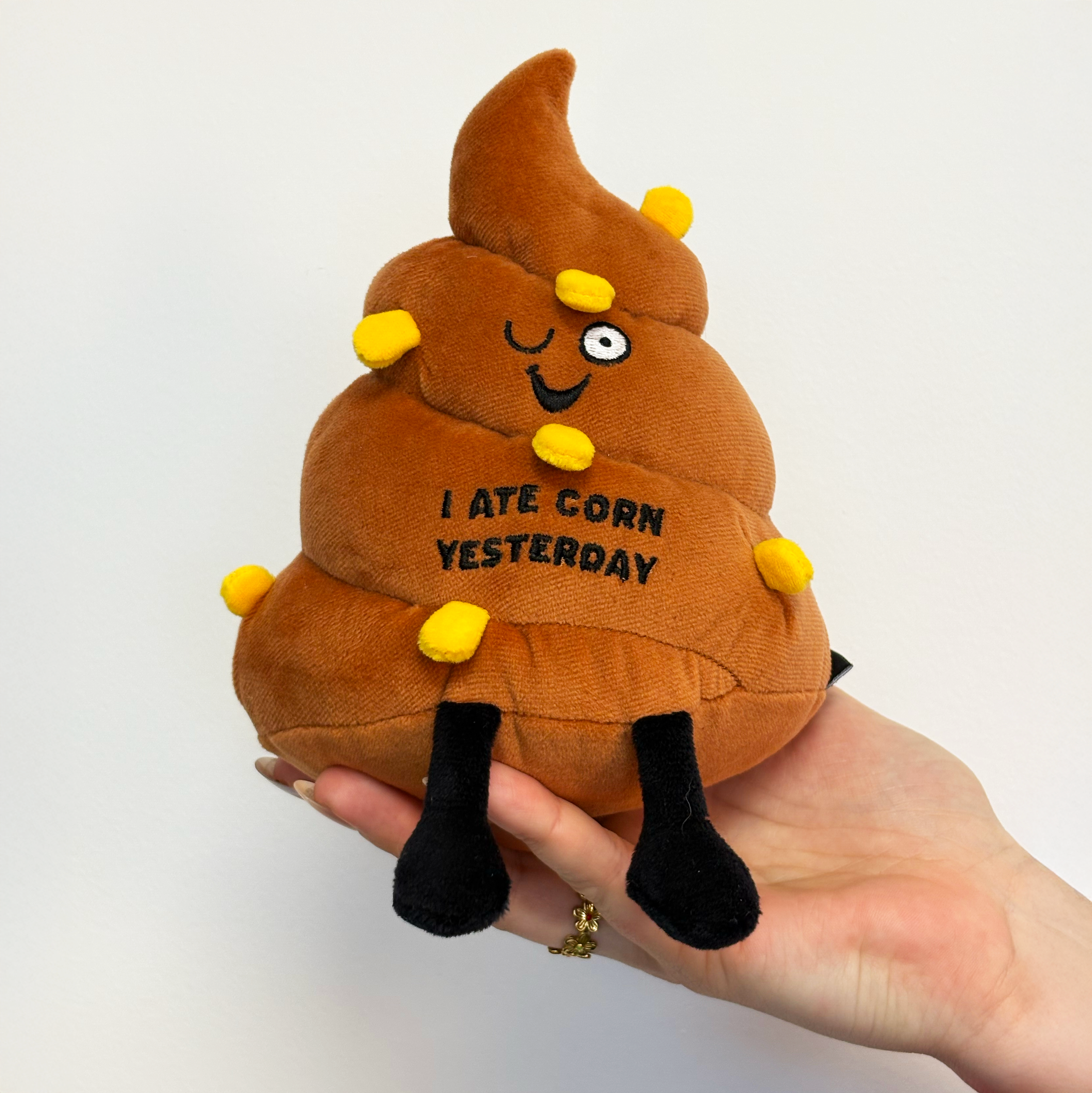 Plush poop with text 'I Ate Corn Yesterday' held by a hand on a white background