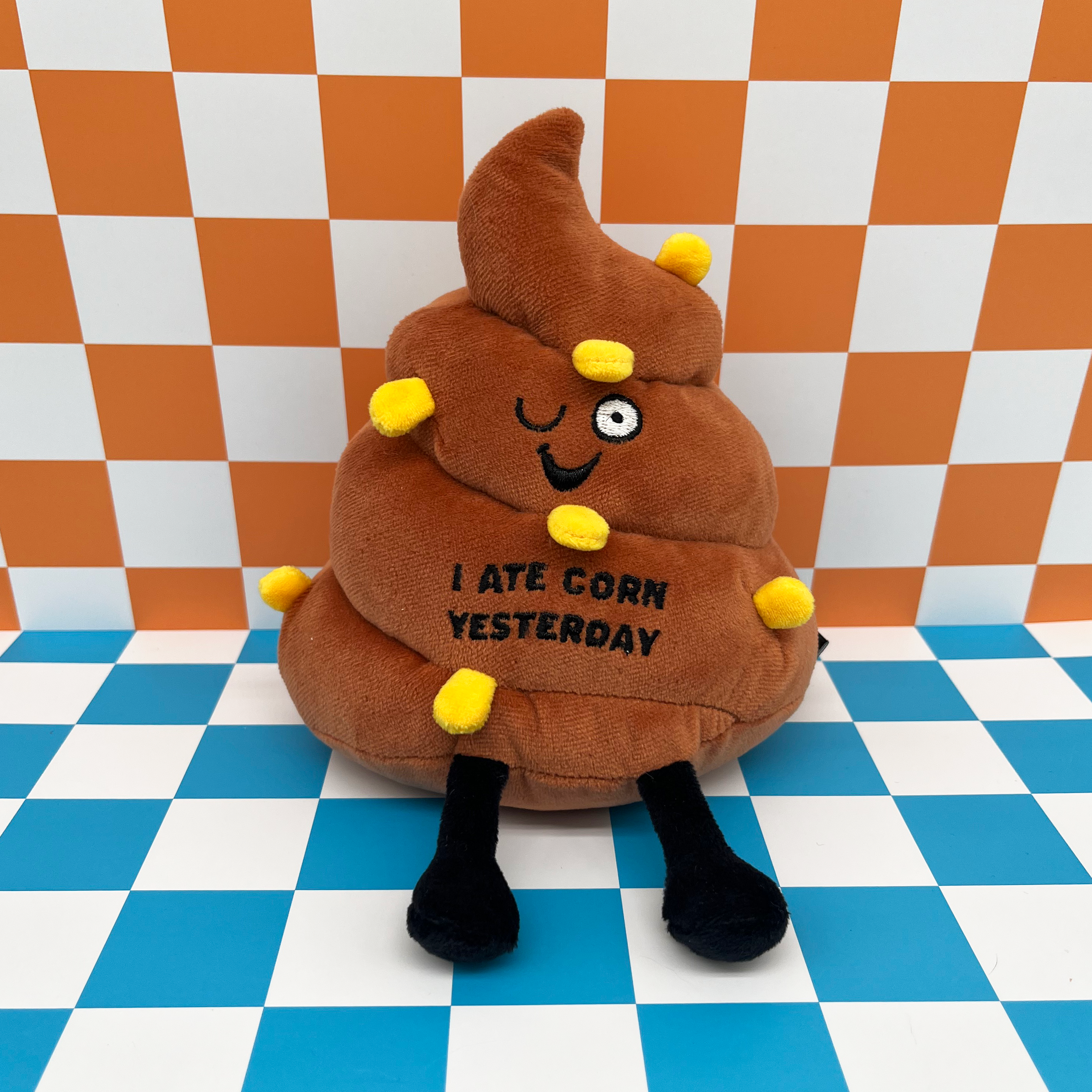 A plush poop with a smiling face, pieces of corn sewn in, and text that reads 'I ATE CORN YESTERDAY' on checkered background. 