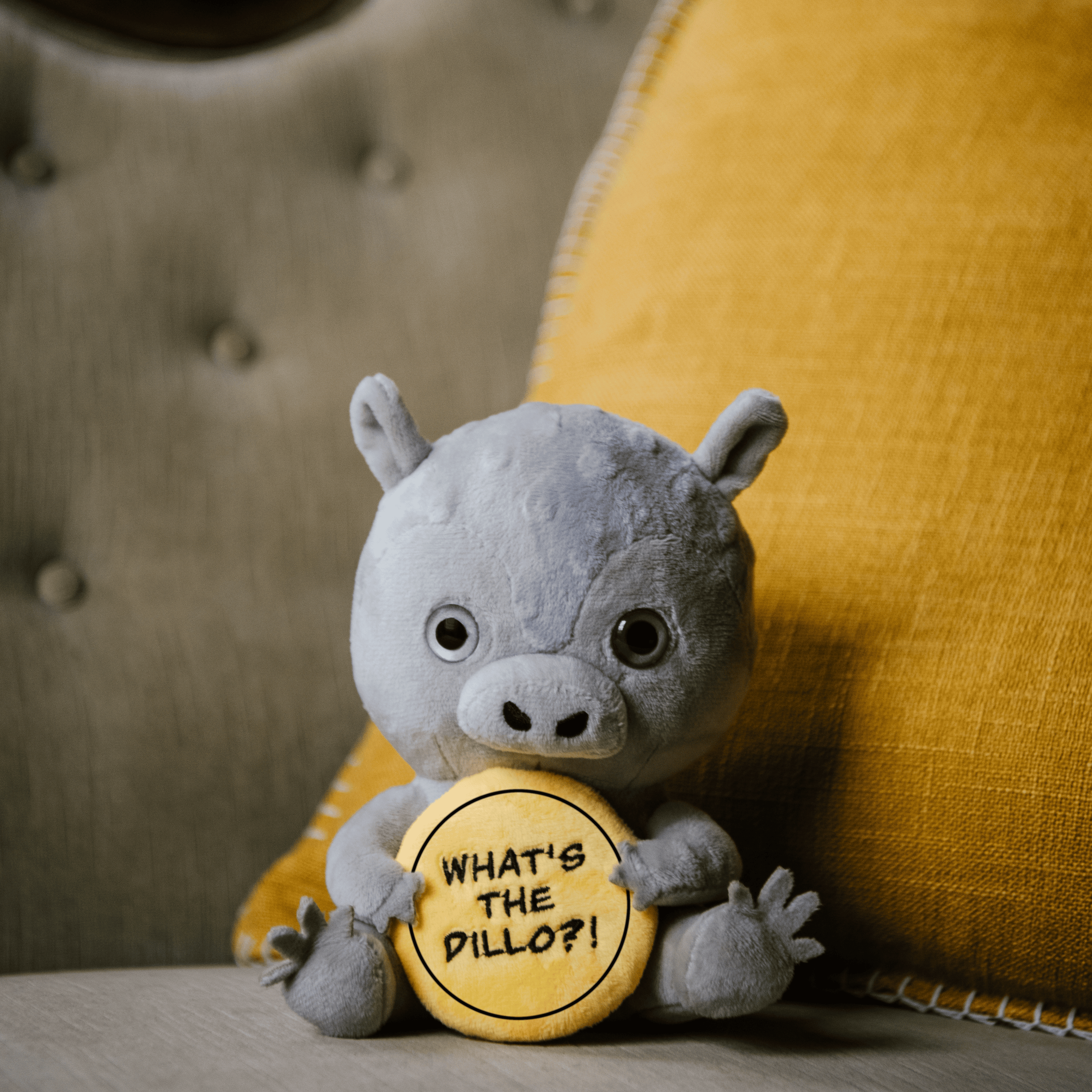 A plush resembling an armadillo holding a yellow sign with the text 'WHAT'S THE DILLO?' placed on a couch.
