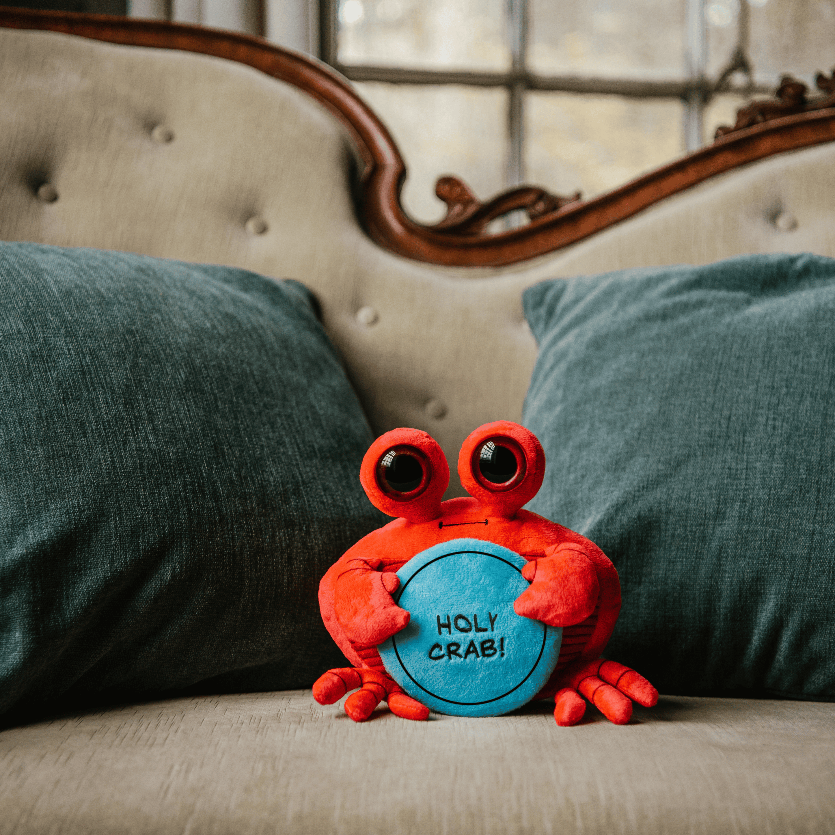 A plush shaped like a crab holding a sign with the text 'HOLY CRAB!' placed on a couch.
